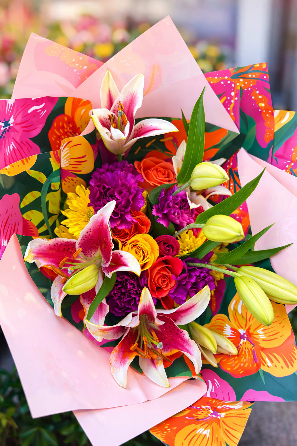 TROPICAL BOUQUET  - Bright pink-and-white lilies take center stage in this tropical flower arrangement, surrounded by fiery orange and red roses, purple carnations, and sunny yellow daisy-like blooms-perfect as a birthday bouquet, congratulations gift, or a cheerful just-because surprise. The vivid mix of saturated warm tones and soft pink accents creates an energetic, celebratory impression that will make the person you're thinking of feel noticed and truly celebrated.  Arranged with layered decorative wrapping-pastel pink paper over a bold tropical floral-print sheet-the presentation reads festive and gift-ready straight from the shop. Textures range from the lilies' broad, silky petals to the ruffled carnations and classic rose blooms; the yellow daisies add light, airy contrast. Wrapped securely for delivery, this arrangement arrives ready to brighten a table or bedside, and will benefit from fresh water on arrival and a cool spot away from direct sunlight to extend its vase life.