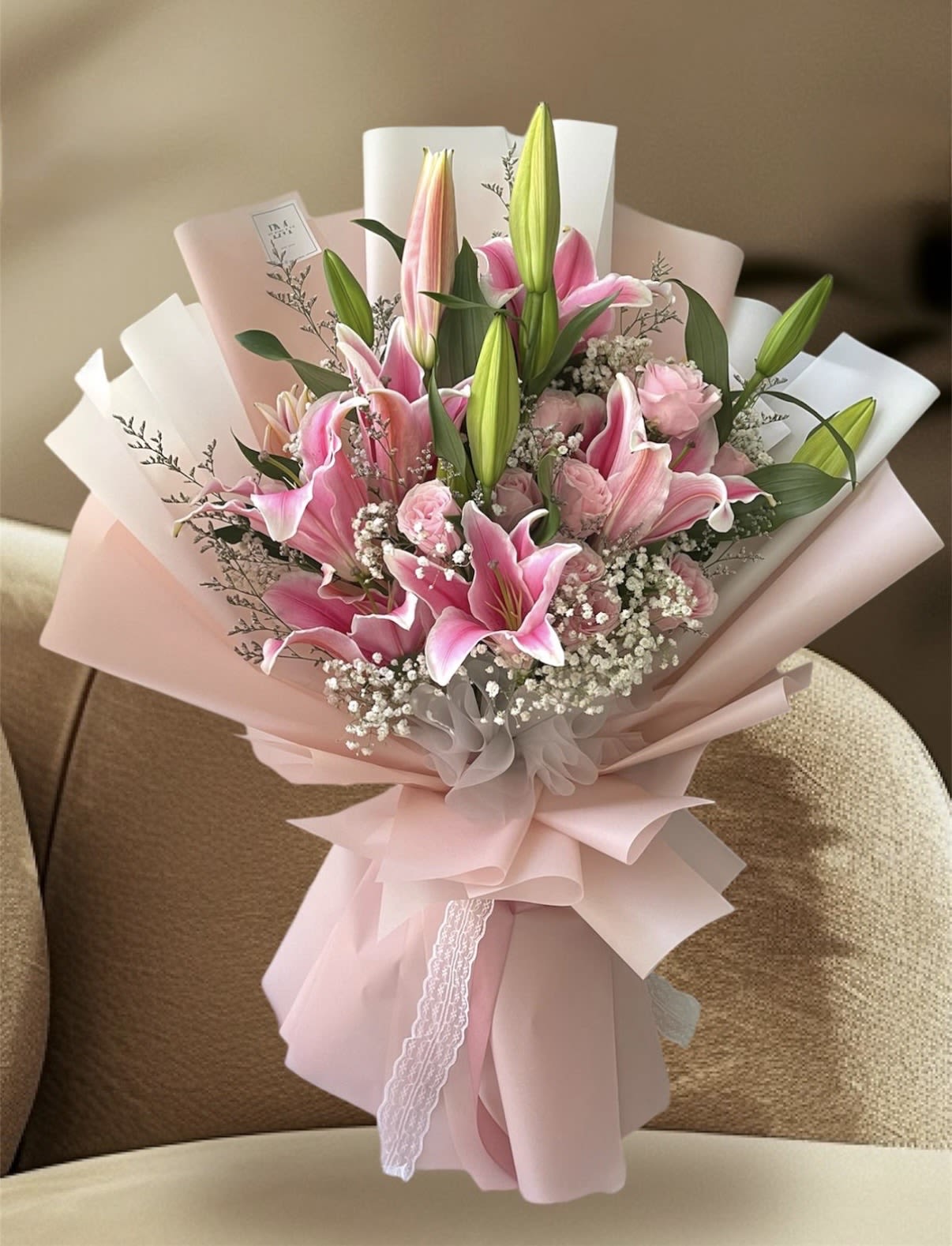 ENCANTO DE LIRIOS  - A cluster of open pink lilies surrounded by soft pink carnations and delicate white filler flowers creates a romantic hand-tied bouquet perfect for an anniversary, Mother's Day, birthday, or a heartfelt thank-you. The upright green lily buds add promise and movement so the person you're thinking of will feel both cherished and pleasantly surprised as blooms open over several days.  Wrapped in layered blush and white paper and finished with a lace ribbon, this arrangement reads feminine and polished-soft textures and a gentle pink, white, and green palette that flatters any setting. The rounded, hand-tied size makes it easy to display in a favorite vase; stems arrive ready to be trimmed and rehydrated so the recipient enjoys lasting fragrance and fresh blooms for days.