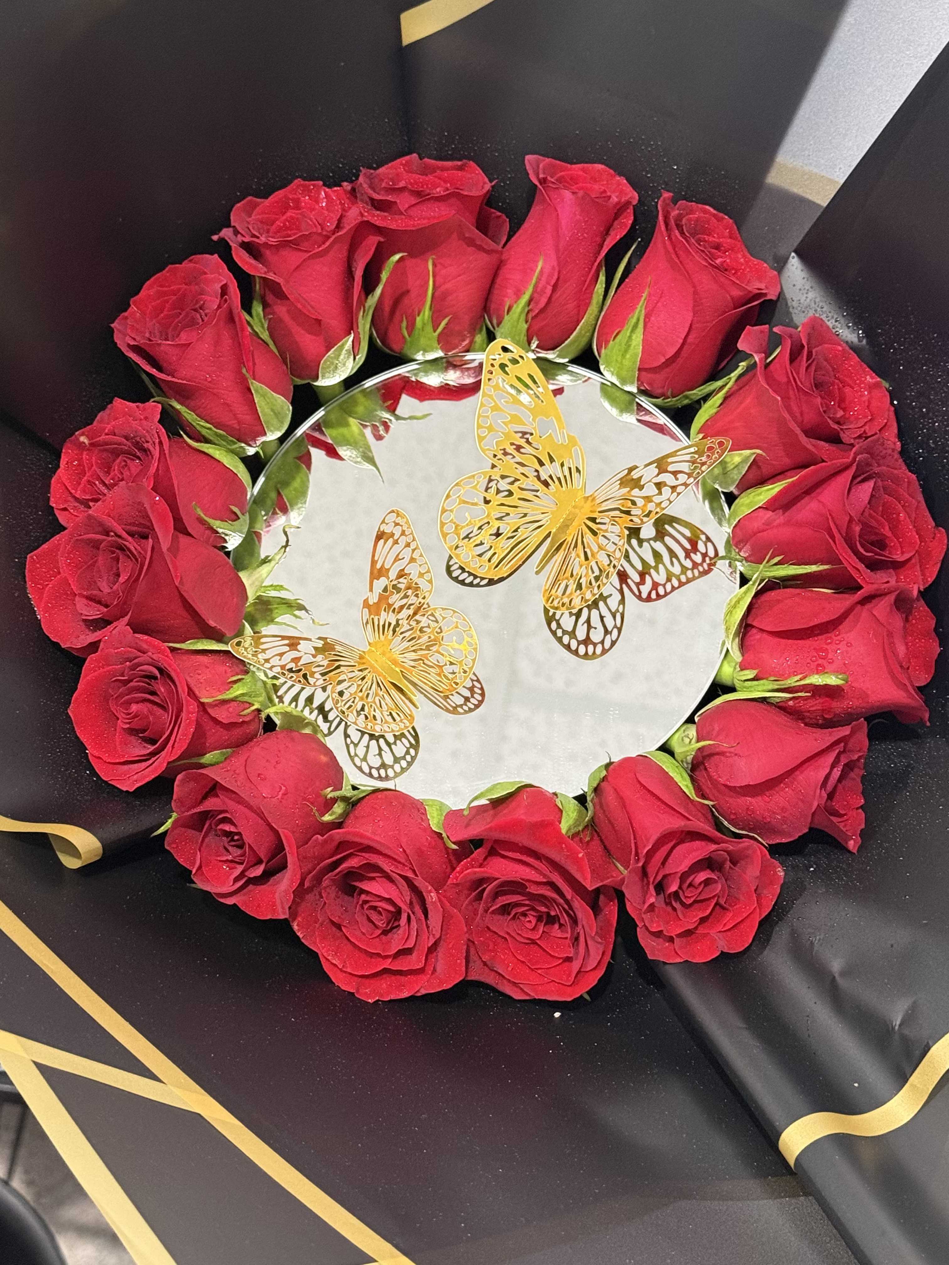 MIR0R BOUQUET - A dramatic ring of red roses encircles a mirrored center in this romantic flower arrangement, perfect as an anniversary bouquet or a bold Valentine's Day gift. The circular design and polished mirror draw the eye inward, making the person you're celebrating feel seen and adored; the red-and-black palette gives the arrangement a passionate, theatrical edge while white and yellow highlights add brightness and contrast.  Tightly packed red roses form the flawless wreath around the mirrored round base, set against black wrapping paper and finished with a gold ribbon for a luxe touch. Two decorative gold butterfly picks rest on the mirror, offering a celebratory sparkle that complements the reflection and heightens the visual impact. The compact, symmetrical shape makes it ideal for tabletop display or a centerpiece where the mirrored surface multiplies the blooms' glow.  This is the kind of arrangement that makes someone feel truly celebrated-suitable for lovers marking a milestone, an elegant birthday surprise, or a memorable proposal setting. Carefully wrapped for gift presentation, the piece arrives ready to place on a table or mantle; keep roses in fresh water after unwrapping and away from direct heat to preserve their vivid color and tight, long-lasting form.