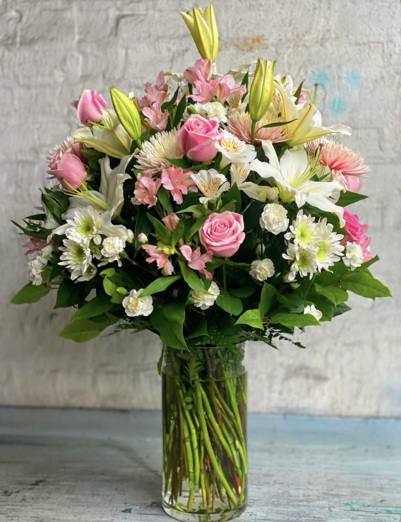 Sweet sensation  - A soft, romantic arrangement that reads like a gentle embrace-Sweet Sensation is a rounded flower arrangement perfect for a birthday bouquet, anniversary flowers, Mother's Day, or a thoughtful thank-you. The mix of pink roses and white lilies creates an immediate impression of tenderness, while bright yellow lily buds lift the composition and add cheerful contrast that catches the eye.  This bouquet pairs pink-and-white alstroemeria, chrysanthemums, and carnations with dense greenery for a lush, layered look. Arranged in a clear glass vase, the design is full and abundant with varied textures-from the velvety rose petals to the trumpet shape of the lilies-so the recipient will feel genuinely celebrated. Vase included; stems arrive arranged and ready to display, with simple care: fresh water and a cool spot will help them stay beautiful longer.
