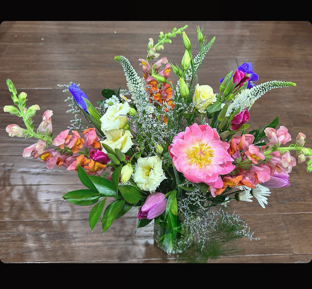 A Walk in the Garden  - A lively, garden-fresh floral arrangement that feels like a sunny spring day   This loose, informal design features lisianthus, snapdragon-like spikes for height and movement, cheerful daisies, and other clustered blooms gathered in a clear glass vase. A large pink focal bloom anchors the front while taller stems and delicate clusters give the arrangement an effortless, natural silhouette. The palette and texture read fresh and uplifting, a walk in the garden.   Sized to sit beautifully on a table or desk, this arrangement arrives ready to display in its glass vase; refresh water and recut stems every few days to extend its life.