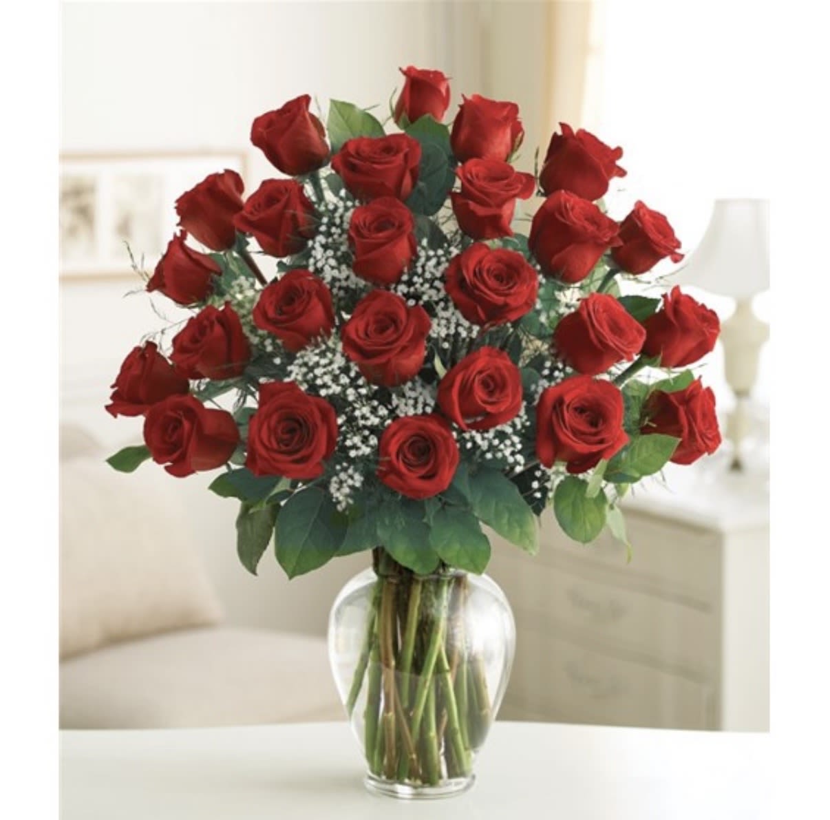 2 DOZEN GLASS - Two dozen red roses arranged in a classic rounded cluster make a bold, romantic statement-perfect as an anniversary bouquet, a Valentine's Day surprise, or a birthday gift that says "you are cherished." The rich red blooms sit upright and close-packed so she'll feel celebrated the moment the vase is set down; delicate white filler flowers and broad green leaves add contrast and depth for an elegant, timeless look.  Presented in a clear glass vase, this arrangement reads as both polished and intimate. The dense grouping of roses creates a full, plush silhouette while the scattered white accents brighten the composition and the green foliage frames the blooms. Sized to showcase two dozen long-stemmed roses, it arrives ready to display and enjoy.  Give this arrangement to the person you're thinking of when you want them to feel unmistakably loved and appreciated; it's the kind of bouquet that marks anniversaries, romantic milestones, or any moment that deserves a heartfelt flourish. For best longevity, place in fresh water, trim stems a bit when you receive it, and refresh the water every few days.