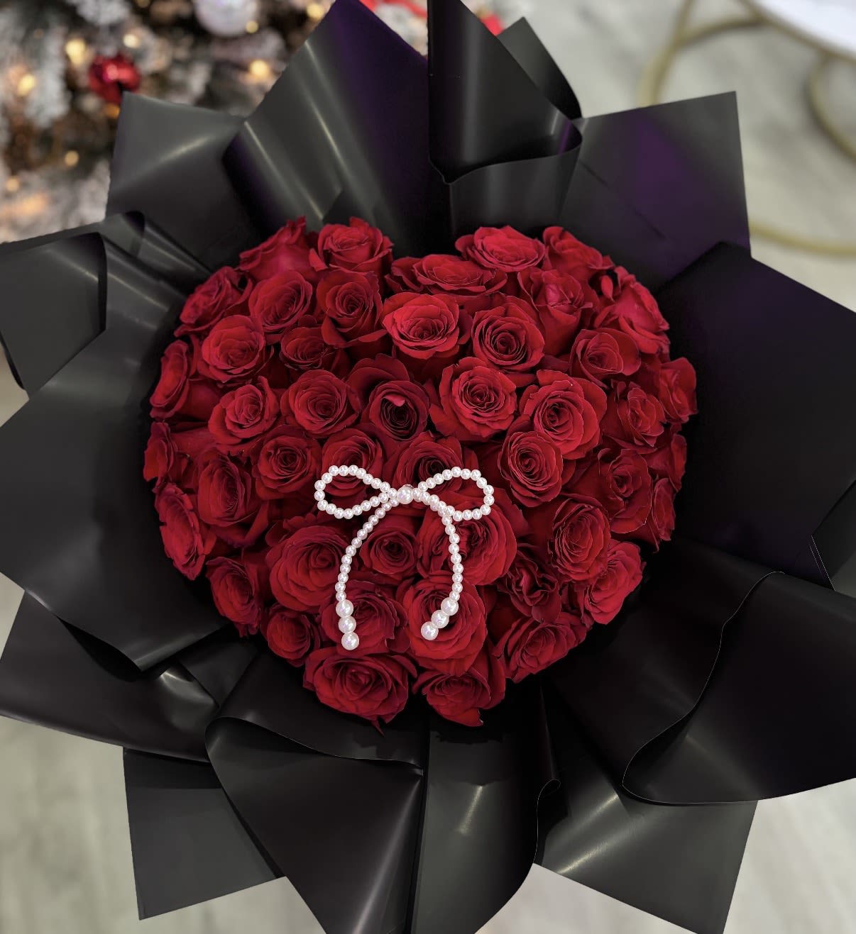 HEART BOCHON  - A dense, rounded bouquet of red roses wrapped in glossy black paper creates a dramatic, romantic statement-perfect as an anniversary bouquet, Valentine's Day gift, or a spontaneous romantic gesture. The striking red-and-black contrast reads bold and sophisticated, the kind of arrangement that makes the person you're thinking of feel deeply noticed and celebrated.  Each bouquet is tightly packed with classic red roses and finished with a small pearl bow at the center for a refined, formal accent. The glossy black wrapping paper adds sleek texture and a modern edge while keeping the focus on the rich crimson blooms. Sized as an everyday gift bouquet, this arrangement is easy to carry and makes an immediate visual impact.  Ideal for partners, spouses, or anyone you want to impress on a romantic occasion, it sends a clear message of passion and devotion. For best enjoyment, keep stems in fresh water, trim stems slightly before placing in a vase, and avoid direct heat to extend bloom life.