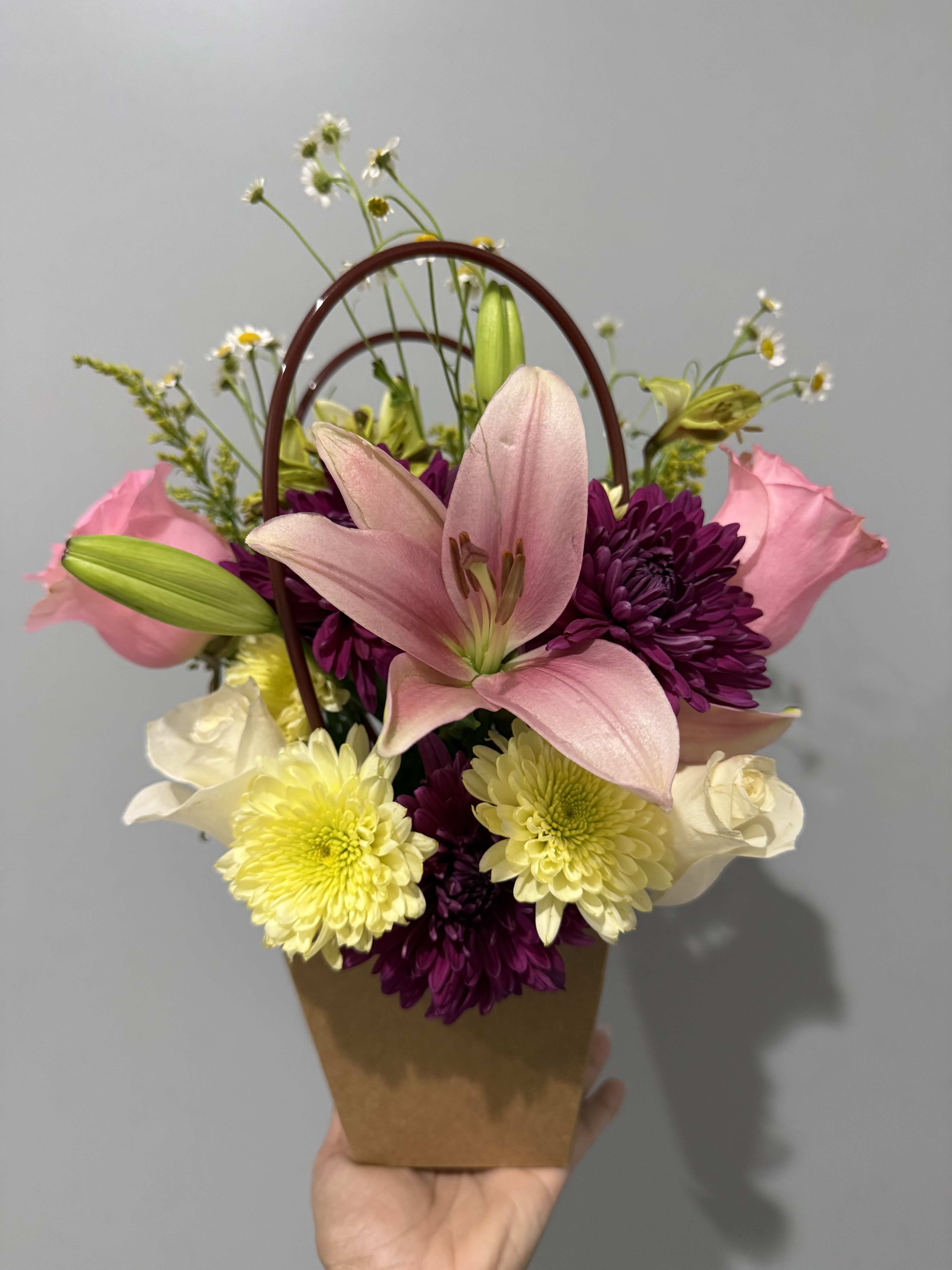 ENCANTO DE PRIMAVERA  - A large pink lily takes center stage in this spring-ready flower arrangement for birthdays, Mother's Day, or a "just because" surprise-the perfect floral gift for someone who loves soft romance with a cheerful twist. The mix of pink roses and deep purple chrysanthemums gives a lush, slightly romantic feel while pale yellow daisy-like blooms and small white filler flowers lift the composition into something bright and airy that will make the recipient feel genuinely celebrated.  Arranged compactly in a brown paper basket with an arched handle, this handheld design reads fresh and informal-easy to carry straight to the table or bedside. The front-facing layout keeps the display neat and focused; expect a tactile contrast between the silky lily petals, velvety roses, textured mums, and daisy centers. Ideal for sending warm wishes or brightening someone's day, this arrangement arrives ready to display with minimal fuss.