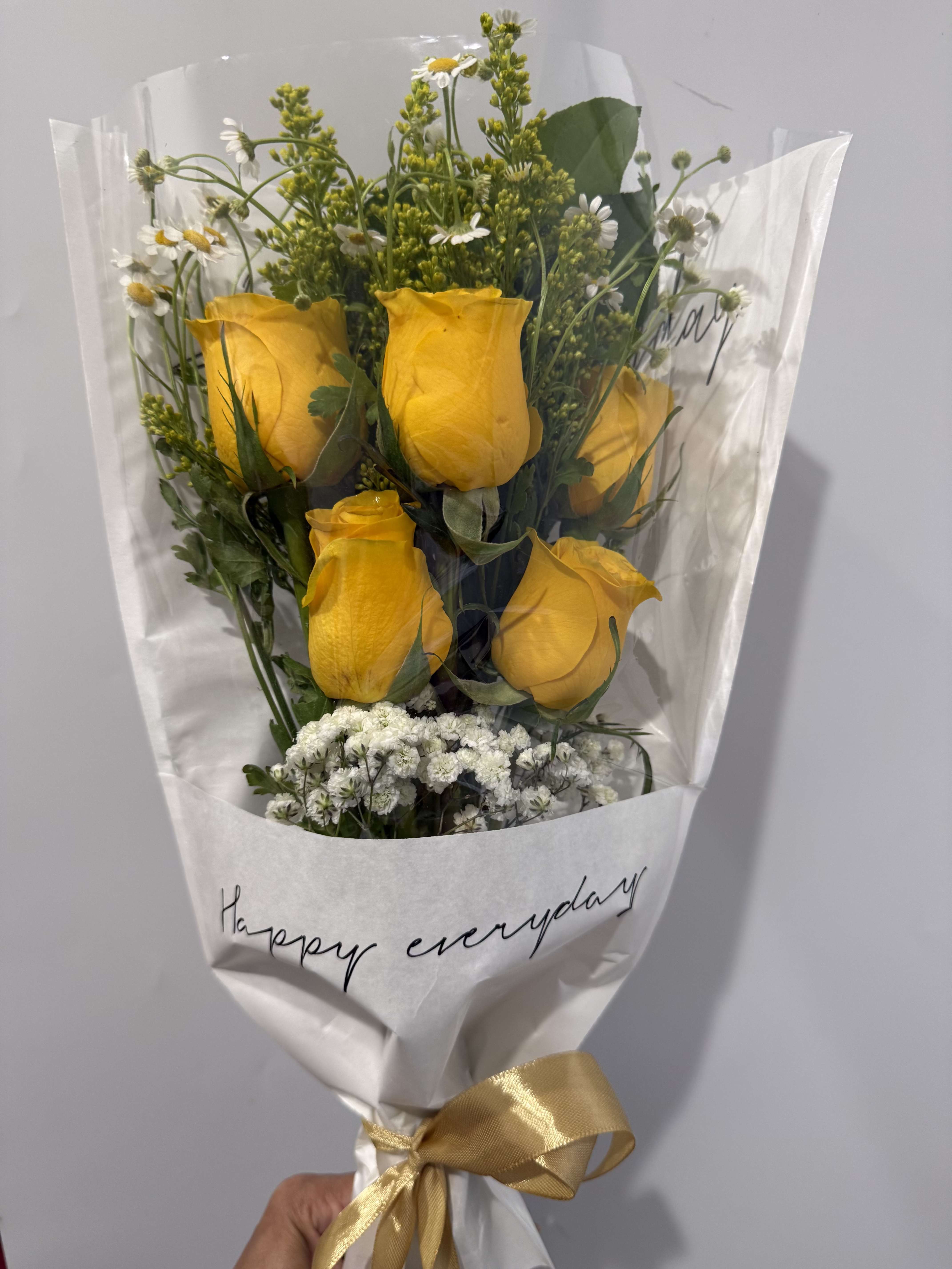 HAPPY EVERYDAY  - Sunny yellow roses paired with breezy white daisies make this everyday bouquet feel like a small celebration - perfect as a birthday bouquet, a cheerful thinking-of-you gift, or a simple way to brighten someone's morning. The loose, hand-tied arrangement reads casual and spontaneous, so the person you're gifting it to will feel genuinely seen and uplifted by its fresh, optimistic color story.  Bright yellow roses sit amid airy white daisy-like filler flowers and fine-textured greenery, wrapped in clear plastic over crisp white paper and finished with a gold ribbon and a handwritten "Happy everyday" note on the wrap. The bouquet's light, approachable scale is ideal for desk or kitchen counter display; stems arrive ready to trim and arrange in a vase, and the open, natural styling keeps the flowers looking lively for days with regular water changes.