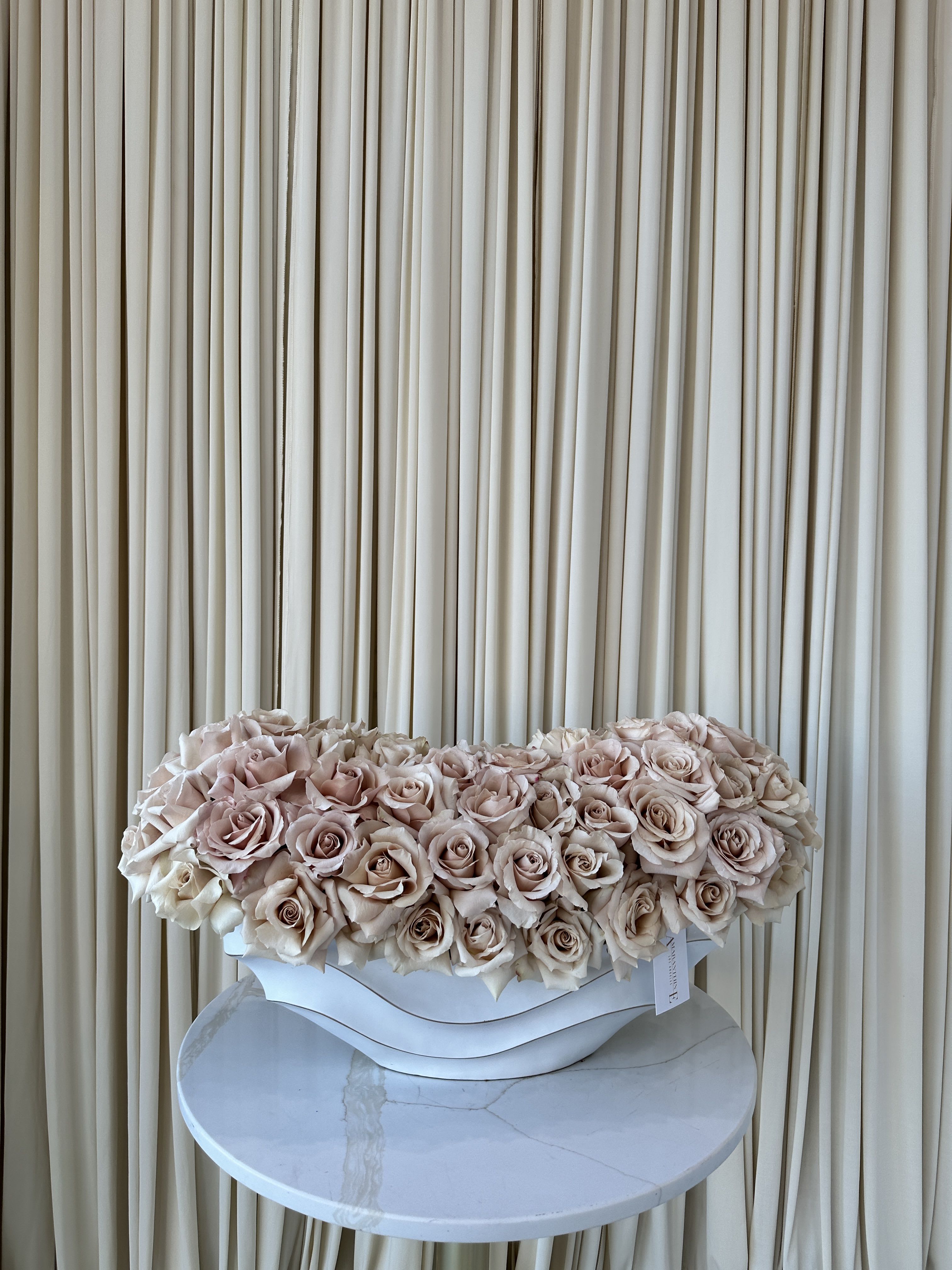 Nude Elegance - A cloud of pale blush and cream roses arranged as a low, luxurious centerpiece-this rose arrangement is a perfect choice for Mother's Day, Valentine's Day, or any special occasion when you want her to feel cherished. The dense, tightly packed blooms form either a broad, horizontal mound or a romantic heart shape, giving a polished, upscale look that reads as both intimate and formal-the kind of gift that makes the recipient feel truly seen.  Each design uses only blush-nude roses in soft pink and ivory tones, placed in a clean white vase.  The texture is velvety and cloudlike, with layered petals creating a full, symmetrical surface ideal for centerpieces, altar displays, or a statement gift on a mantel.   Made to convey tenderness and timeless elegance, this arrangement is especially suited for mothers, partners, or anyone you want to honor on a milestone or intimate celebration. Care is simple: keep out of direct sun for longest vase life; order early for holiday deliveries to ensure availability.
