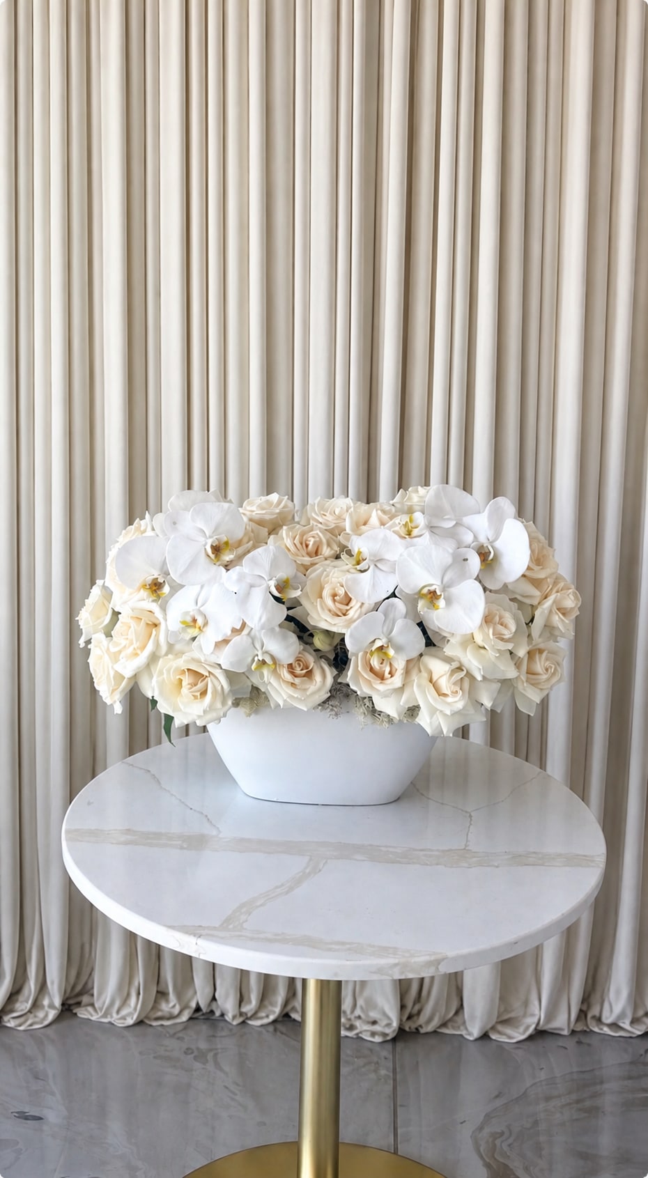 Blooming Orchids - A low, luminous cluster of cream and white roses topped with large white orchids gives this flower arrangement an elegant, serene presence-perfect as a wedding centerpiece, anniversary gift, or refined everyday surprise. The compact, symmetrical mound reads as calm and polished, the kind of floral statement that will make the person you're thinking of feel truly celebrated and seen.  Tightly packed cream and white roses form a soft base, while the glossy white orchids arch gracefully above, adding sculptural contrast and a subtle perfume. The blooms are arranged in a smooth white ceramic vase that emphasizes the pale, sophisticated palette.  This arrangement suits couples marking a milestone, hosts seeking an elegant table accent, or anyone who appreciates refined, low-profile florals. It arrives ready to display in its white ceramic vessel-minimal care like fresh water and a cool spot will keep the blooms at their best-making it an effortless, memorable gift.
