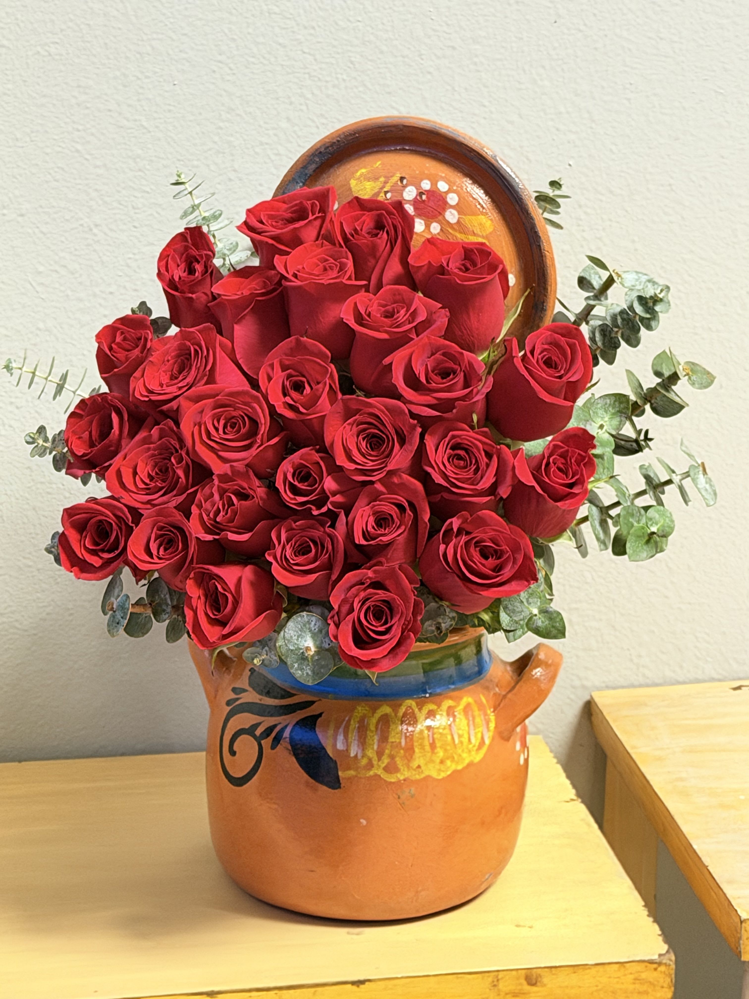 Ollita perfecta  - A dense cluster of red roses gathered into a rounded bouquet creates an immediate impression of warmth and devotion - an ideal Mother's Day arr or anniversary roses to make the mother in your life feel adored. The bright red blooms read as classic romance while the orange ceramic pot with painted decorative details gives the gift a handcrafted, artisanal charm that makes her feel specially noticed.  The arrangement sits low and full in the ceramic pot, the painted finish adding a pop of color and a tactile, homey quality. Eucalyptus-like green foliage frames the roses, softening the intense red and adding subtle texture and movement; the red, orange, and green palette feels festive and celebratory without being fussy.  Choose this for Mother's Day, an intimate celebration, or to say "thank you" in a way that honors her presence. The ceramic pot arrives ready to display and becomes part of the gift; light trimming of foliage and fresh water will keep the roses looking their best for days.ngment 