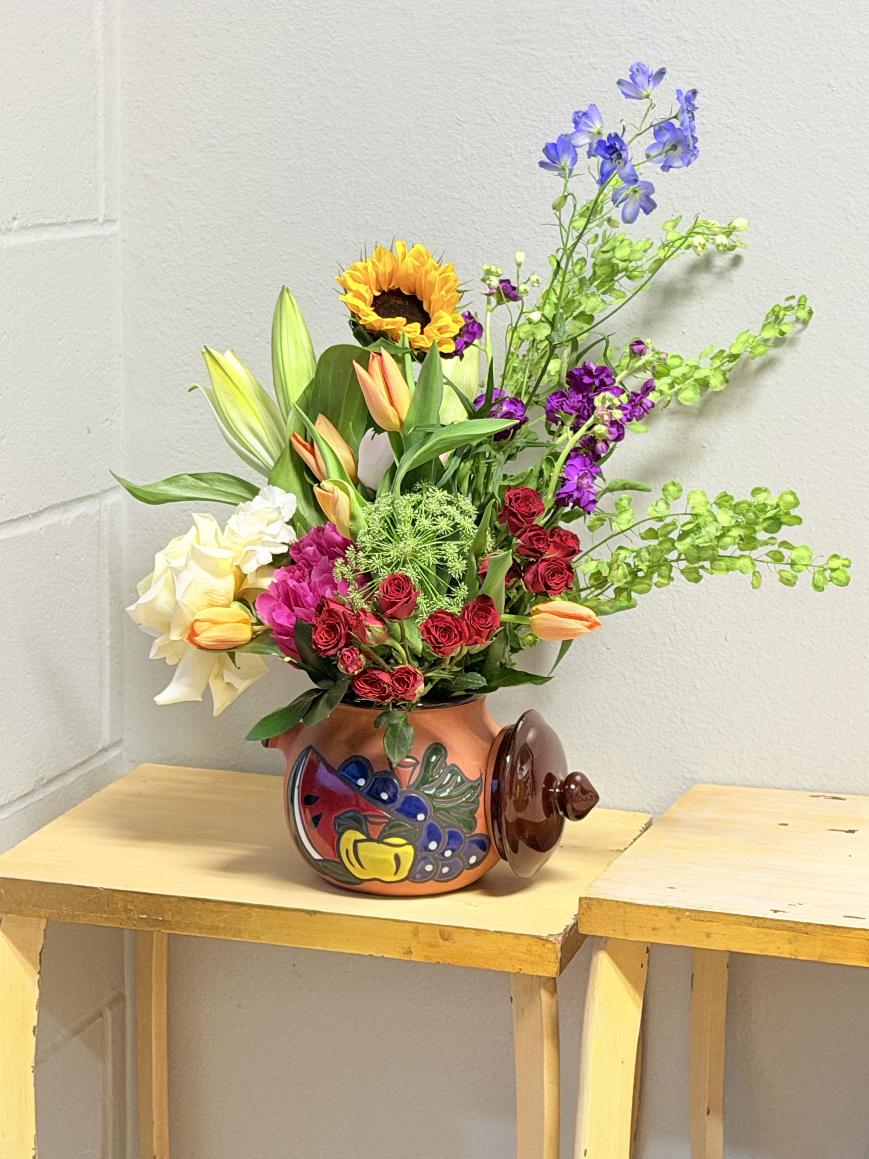 Fruitie Cutie - A bright, joyful centerpiece that reads like a sunlit garden-this flower arrangement is a cheerful mixed-color bouquet perfect as a birthday bouquet, Mother's Day gift, or thank-you arrangement. Sunflowers, white lilies, tulips in peach and white, and red spray roses create a warm, celebratory palette, while tall blue-green stems and delphinium-like spikes give the piece lively height and a loose, asymmetrical silhouette that feels festive and spontaneous. The overall effect is the kind of bouquet that makes the person you're thinking of feel truly seen and celebrated.  Carefully arranged in a painted ceramic or painted terracotta vase with folk-art fruit motifs and a lid-like side detail, the design blends compact abundance with upward movement-bright yellows and oranges contrast with cool blues and purples for extra visual pop. Accents of stock and other seasonal blooms fill the composition for texture and fragrance; on the Mother's Day menu this style includes a sprinkle of peonies and exclusive blooms for added softness. Sized to brighten a table or entryway, it arrives ready to display-trim stems and refresh water as needed to extend its lively, gift-ready presence.