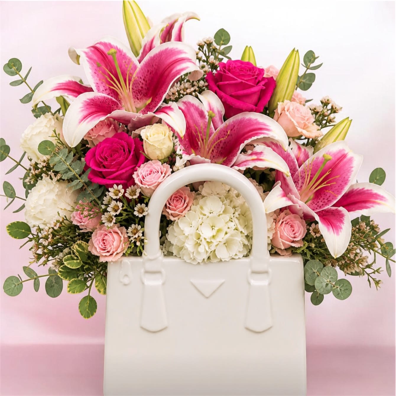 Elegant Bloom Purse - A stunning expression of elegance and modern floral design, the Elegant Stargazer Purse Arrangement is crafted in a unique handbag-shaped ceramic vase that adds a sophisticated and luxurious touch. This exquisite arrangement features fragrant stargazer lilies, premium roses in shades of pink, delicate spray roses, and lush white hydrangeas, beautifully complemented with fresh eucalyptus and fine fillers. Each bloom is carefully placed to create a harmonious, full, and eye-catching composition. Perfect for birthdays, anniversaries, celebrations, or simply to make someone feel truly special, this design blends romance, style, and creativity in one unforgettable gift.  A statement piece that transforms flowers into art.