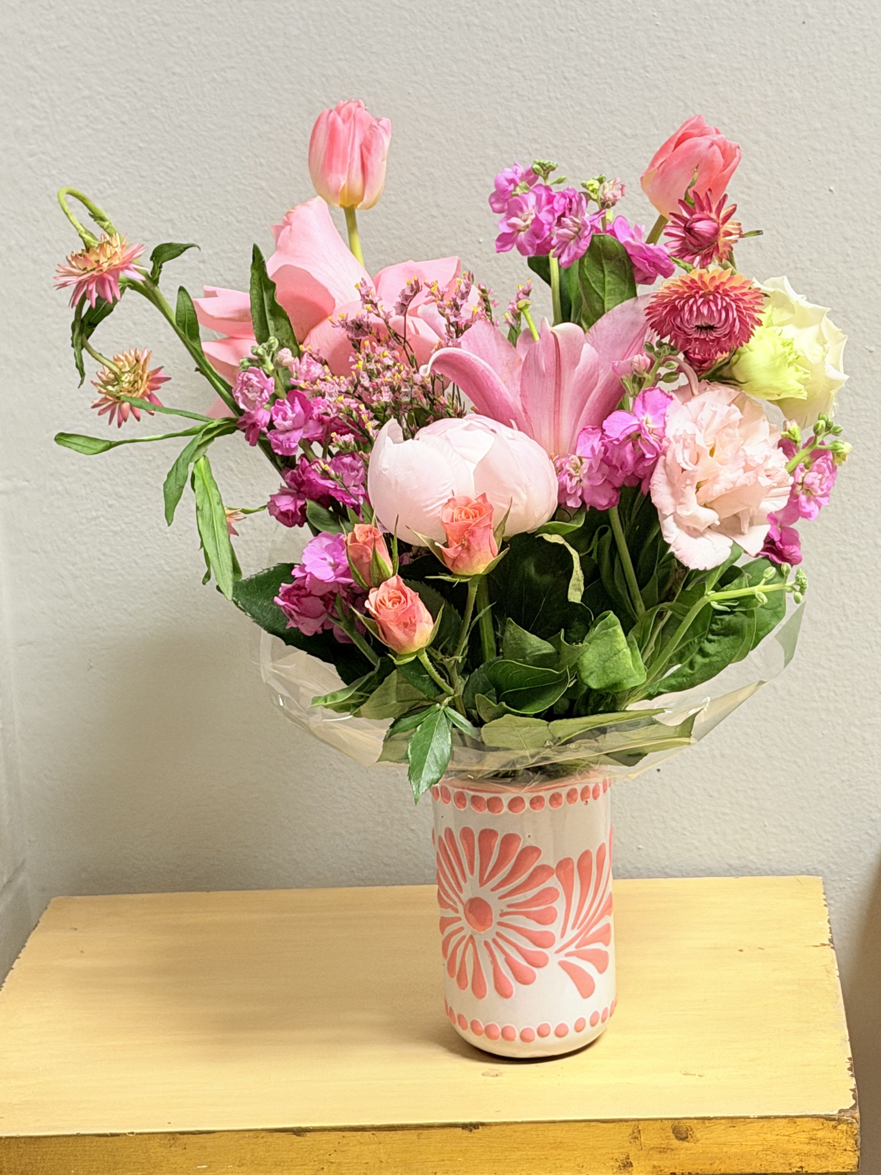 Pink floresitas  - A lively mix of pinks and soft whites makes this flower arrangement an instantly cheerful birthday or "just because" gift that will brighten a Mother's Day table or any spring celebration. The upright bouquet of tulips, a large pink lily, roses, carnations, lisianthus, peonies and stock creates a romantic, lightly fragrant display that reads fresh and joyful - the kind of arrangement that will make the person you're thinking of feel truly celebrated.  Arranged loosely for a natural, garden-picked look, the blooms sit in a white ceramic vase with pink floral patterns and are wrapped in clear plastic for delivery. The palette combines vivid and pastel pinks with white accents and pops of bright green foliage; textures range from the silky tulip petals to the ruffled carnations and clustered filler flowers. Dimensions and stem count vary by size selected; vase is included and ready to display.  Perfect for springtime gifting, birthdays, or sending a spontaneous note of affection, this arrangement needs only fresh water and a cool spot away from direct sun to keep blooms at their best. Order with confidence for a recipient who loves romantic pinks and a handmade, decorative touch.