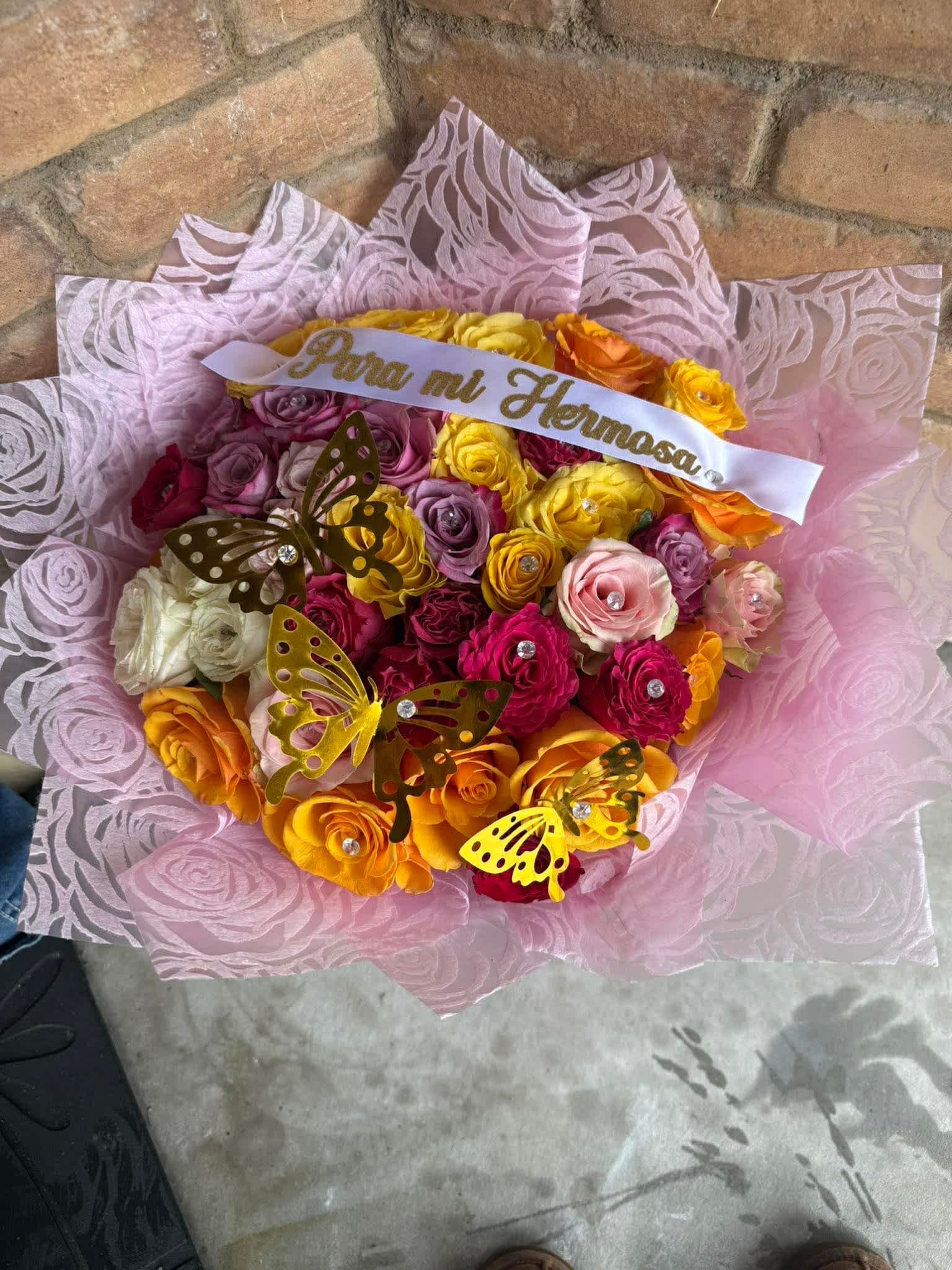 Ramo buchon rainbow  - A tightly packed celebration of color, this rose bouquet is a joyful birthday bouquet and romantic gift all in one-perfect for saying "feliz cumpleaños," "te quiero," or simply brightening someone's day. The mix of pink, yellow, orange, red (with touches of lavender and white) creates a playful, uplifting palette that reads as both festive and affectionate, so the person you're thinking of will feel celebrated and adored the moment they see it.  Each bouquet features a generous cluster of roses arranged close together and wrapped in layered pink patterned paper for a luxe, present-like look. A white satin ribbon printed with gold "Para mi Hermosa" lettering finishes the package, while gold butterfly decorations and small rhinestone embellishments give the design a sparkling, whimsical touch. The combination of warm rose tones and embellished hard goods makes this arrangement especially well suited for birthdays, anniversaries, or any celebratory moment when you want her to feel truly seen.  This is the kind of bouquet that reads as a keepsake as well as flowers-no extra vase required if you want to present it as a gift; the wrapping and ribbon make unwrapping part of the experience. For best enjoyment, place the roses in fresh water as soon as possible and trim stems before arranging to extend vase life; it's an immediate, joyful gift that delivers a clear message of affection and celebration.