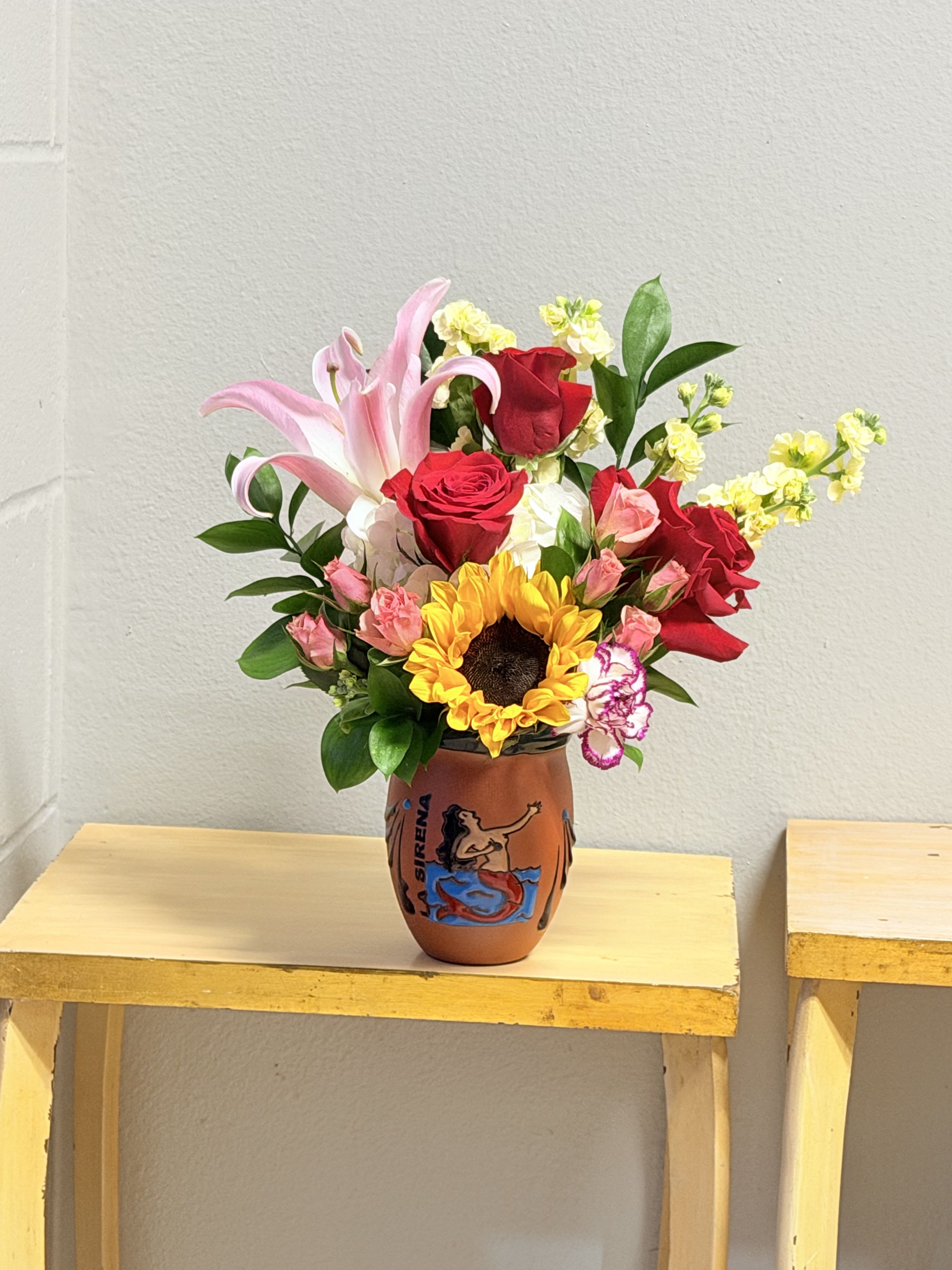 La sirena  - A single bold sunflower centerstage brightens this mixed bouquet, surrounded by red and pink roses for a touch of romance-perfect as a birthday bouquet, thank-you gift, or just-because surprise. The combination of warm reds, soft pinks and vivid yellow reads cheerful and lively, a floral way to make the person you're thinking of feel noticed and celebrated.  This arrangement pairs a pale pink lily and pink carnations with small yellow filler blooms and other seasonal greens, all gathered in a hand-painted ceramic vase that adds a crafted, decorative touch. The vase gives the display weight and personality while the mix of textures-from the sunflower's bold face to the roses' layered petals-creates visual movement and long-lasting presence.  Made to suit many occasions, this bouquet works for a friend, mother, partner, or colleague who appreciates a bright, slightly romantic statement. Vase included; enjoy regular fresh water changes and trim stems if you'd like to extend vase life.
