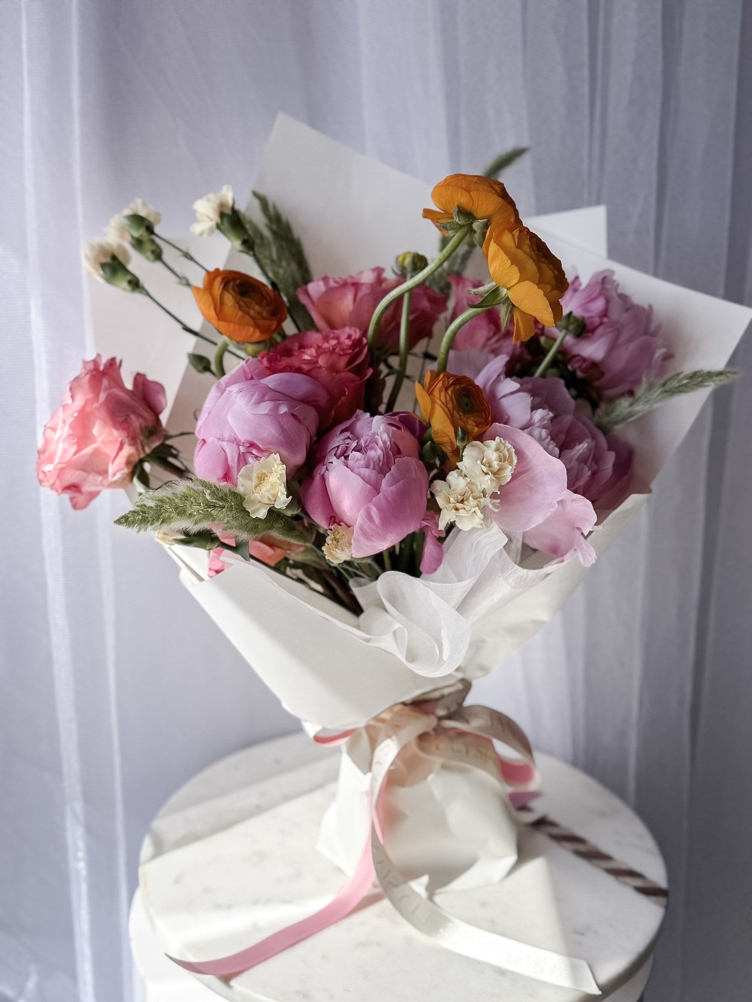Peach Blossom - A joyful, garden-inspired bouquet that feels like a warm breeze on a spring morning. Peach Blossom brings together lush pink peonies, playful orange ranunculus, and delicate accents in soft cream tones, creating a vibrant yet romantic palette. Wrapped in crisp white with flowing ribbons, it captures a perfect balance of elegance and spontaneity.