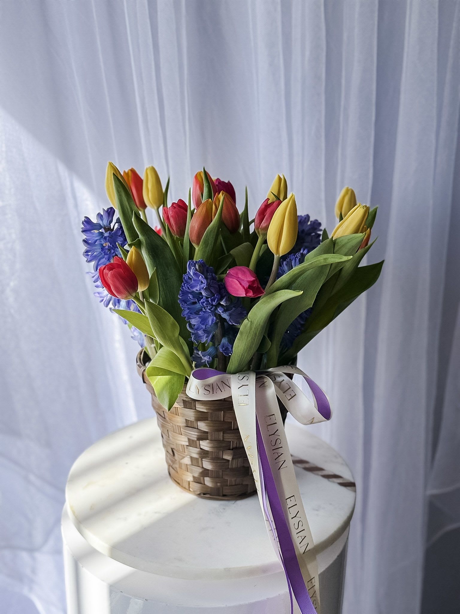 Spring Melody Basket - A cheerful symphony of color and texture, Spring Melody Basket captures the essence of the season in full bloom. Featuring vibrant tulips in sunny yellows, warm oranges, and playful pinks, paired with fragrant blue hyacinths, this arrangement bursts with life and freshness. Nestled in a charming woven basket and finished with soft, flowing ribbons, it feels both inviting and thoughtfully curated.