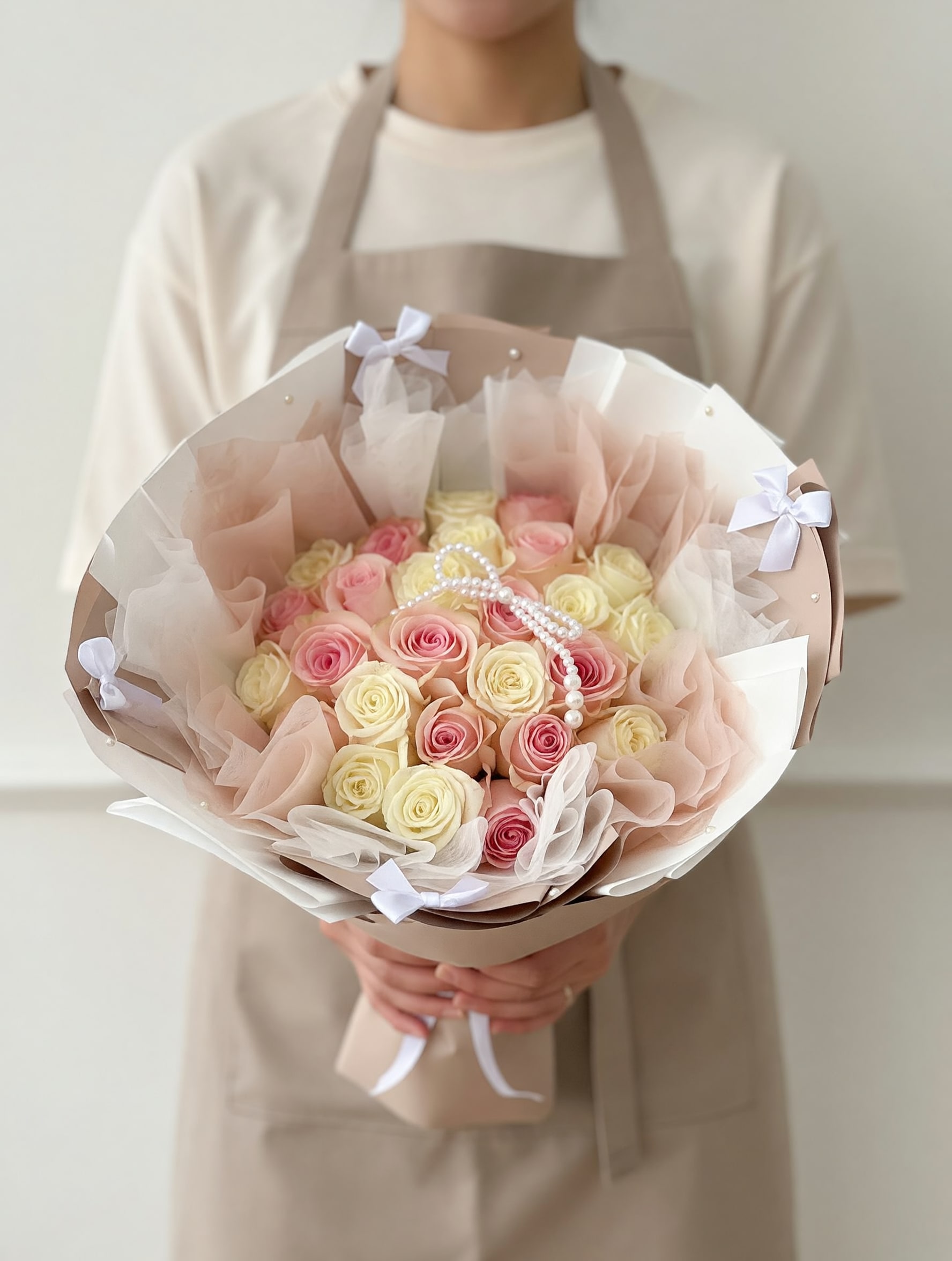 Pearl Blush Rose Bouquet - A luxurious bouquet featuring a delicate blend of blush pink and ivory roses, beautifully wrapped in soft layered paper and adorned with elegant ribbon details and a pearl accent. Pearl Blush Rose Bouquet is designed to express pure love, elegance, and timeless beauty—perfect for unforgettable moments.