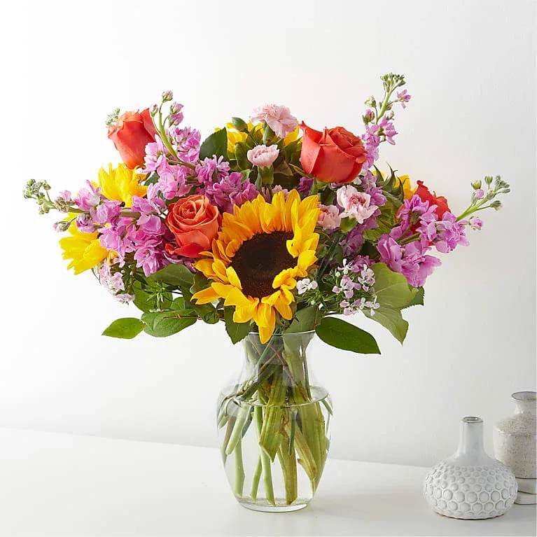 Yellow Brick Road Bouquet - Bright, sunny sunflowers take center stage in this yellow, pink, orange and white flower arrangement-an uplifting birthday bouquet or thank-you gift that immediately brightens a room. The Yellow Brick Road Bouquet reads like a cheerful walk through summer: bold yellow and warm orange tones deliver instant optimism while pink accents add a soft, friendly note that will make the person you're thinking of feel genuinely celebrated.  The mix includes large sunflowers, orange roses, carnations and clusters of pink and lavender filler blooms arranged in a clear glass vase so stems are visible through the water for a fresh, casual look. The palette balances high-impact color with delicate texture; expect a full, hand-tied style approximately vase-height, perfect for a mantel, kitchen table, or desk.  Ideal for birthdays, thank-yous, or any spring and summer moment when you want to lift someone's spirits, this arrangement arrives ready to display-no extra styling needed. To keep the bouquet looking its best, refresh the water every couple of days and trim stems if desired before placing back in the vase.