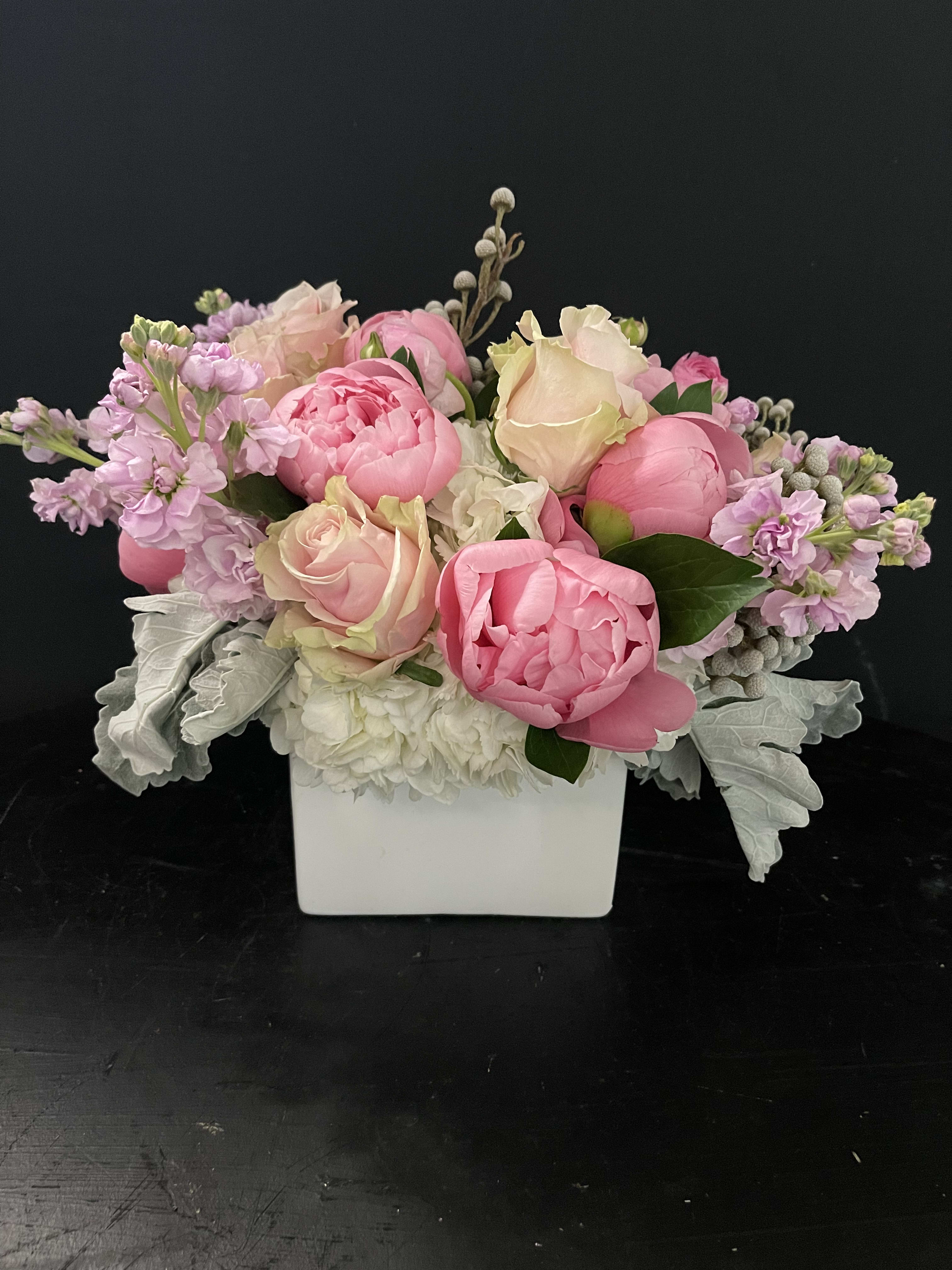 Low arrangement of pink peonies and blush roses in a white square vase with soft white flowers.