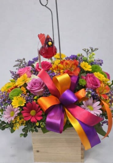 “Enchanted Love Garden”  by Art and Flowers - This arrangement bursts with cheerful, high‑energy color — it feels like a celebration captured in flowers.  A lively mix of blooms fills a light wooden box, creating a bright, joyful display. Pink roses add softness, while yellow daisies and green button mums bring a sunny, playful vibe. Purple asters and magenta blossoms deepen the palette with rich contrast. The oversized bow — a swirl of orange, purple, and hot pink ribbons — gives the whole piece a festive, gift‑like personality. We will vary   Hovering above it all, a small red glass bird perched on a metal hook adds a whimsical, almost storybook charm, as if it’s watching over the garden below.  We will vary the blooms and colors based upon availability and seasonality. 