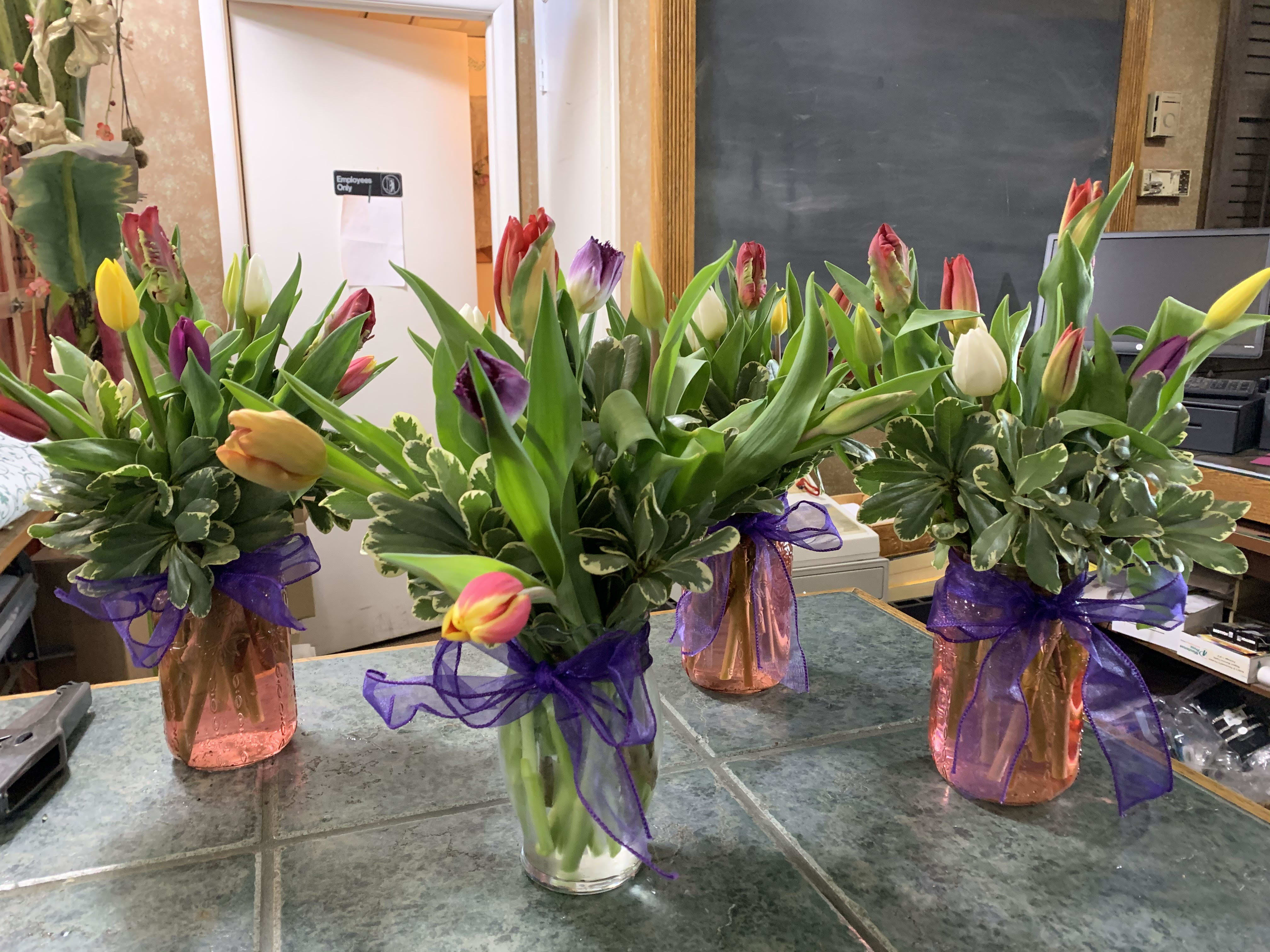 Four arrangements of mixed-color tulips in small glass vases with purple ribbon bows.