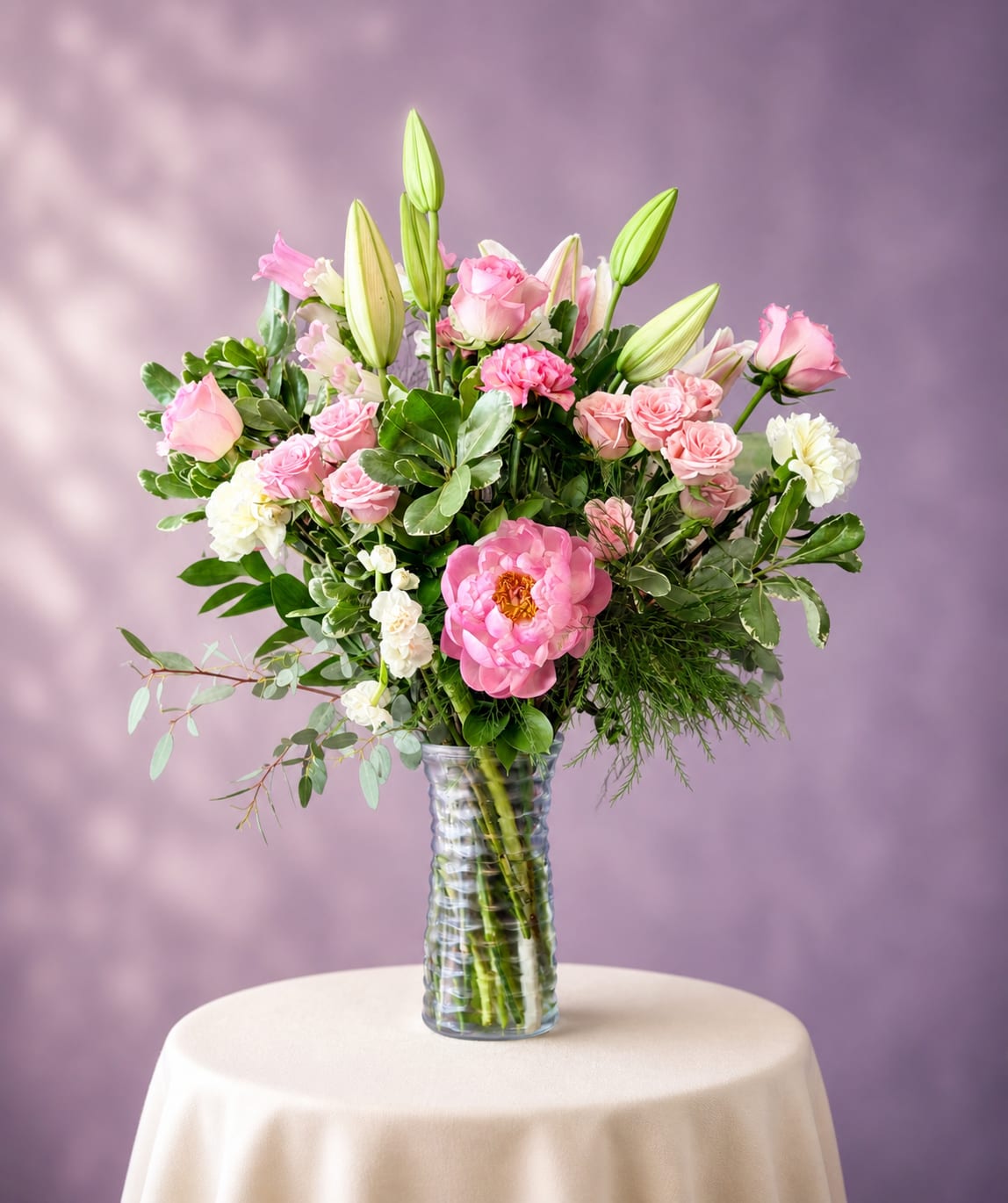 Mother Day Special  - Soft pink roses and lush garden-style blooms make this Mother's Day floral arrangement an instant favorite for celebrating mom, birthdays, anniversaries, or simply saying "I'm thinking of you." The upright, full design and feminine palette of pink, white, green and a hint of purple creates a romantic, comforting impression that will make the person you're thinking of feel cherished and remembered.  A generous mix of pink roses, large pink peony-like blooms, white carnations and unopened lily buds are artfully arranged with fresh green foliage in a clear ribbed glass vase. The bouquet spreads wide at the top with a balanced, natural silhouette that narrows toward the vase, giving a lush, hand-gathered look. The clear glass allows stems to show through for a clean, classic presentation that suits tabletops, mantels, or bedside tables.  Perfect for Mother's Day or any occasion that calls for warmth and tenderness, this arrangement reads as both celebratory and sincere-the kind of gift that makes her feel truly seen. Vase included; water upon delivery and trim stems every few days to keep blooms fresh longer.