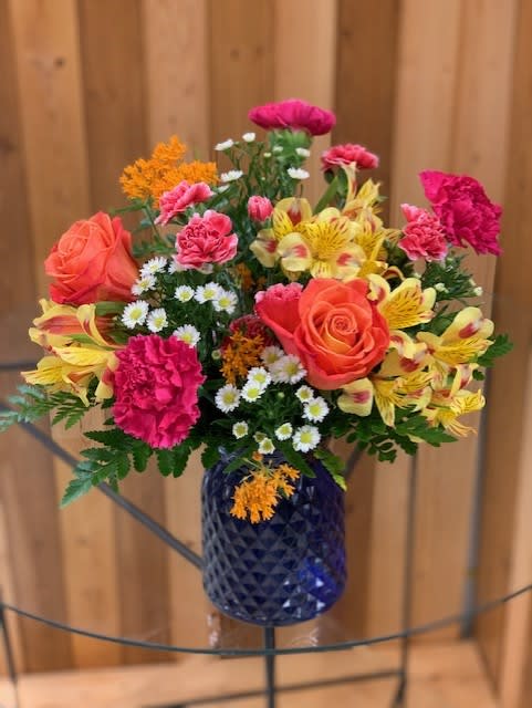 Bright mix of orange roses, pink carnations, yellow alstroemeria, and daisies in a blue glass vase.