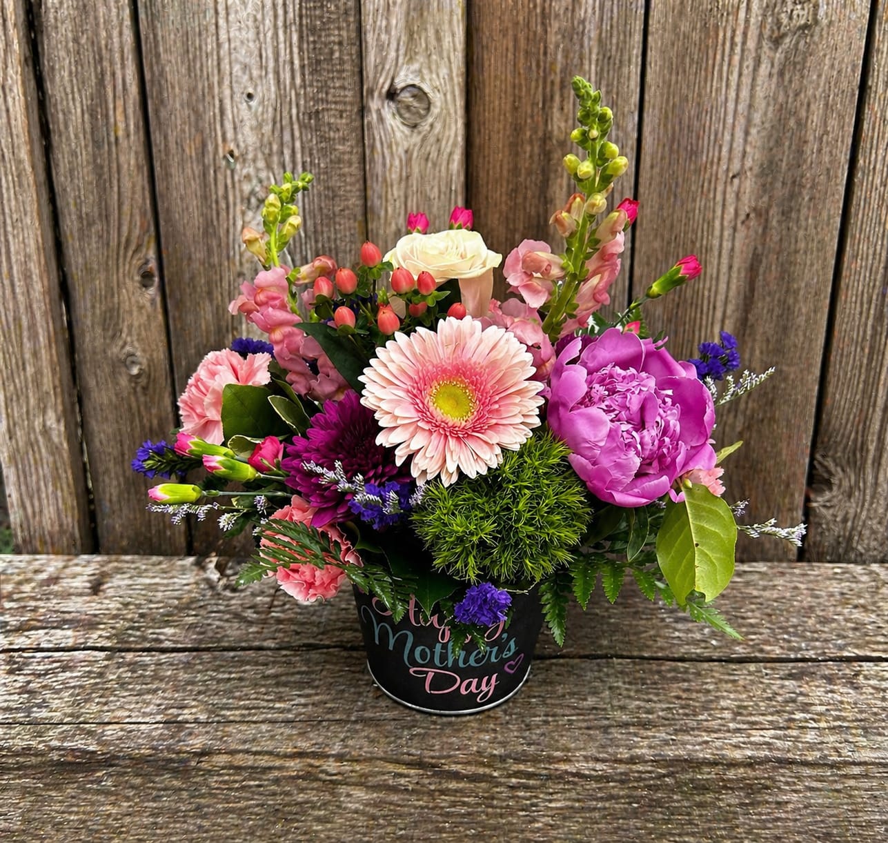 Mother's Spring Garden - A bright, compact spring bouquet made for Mom: soft pink gerbera daisies and a bold purple peony-style bloom anchor this rounded Mother's Day arrangement, surrounded by coral and white carnations, snapdragons, and cheerful daisies. The mix of pink, purple, white, and green creates a feminine, uplifting palette that reads as both celebratory and comforting-perfect as a Mother's Day gift or a springtime surprise to make her smile.  Arranged densely in a black decorative container printed with "Mother's Day" lettering, the design feels full and abundant while remaining neatly contained. Snapdragons and smaller filler blooms add vertical movement and contrast to the round silhouettes of the gerbera and peony-like flowers. The compact size is easy to display on a table or counter; stems arrive arranged for immediate enjoyment with standard floral care-fresh water and a trimmed stem if needed.