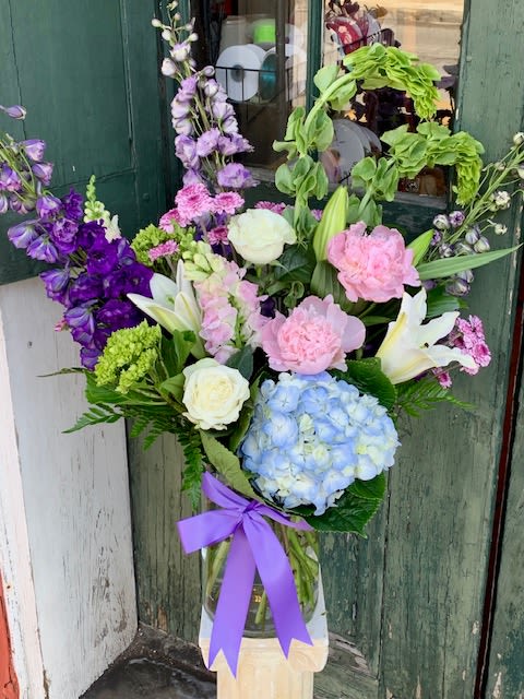 Mother's Day 2025 - A tall, garden-style flower arrangement that feels like a springtime hug-perfect as a Mother's Day bouquet or for a birthday, anniversary, thank-you, or romantic surprise. The centerpiece is a large blue hydrangea set against creamy white lilies and roses, with soft pink peonies or carnations and vertical purple snapdragon-like spikes for height. The overall palette of blue, lavender, pink, white and purple reads feminine and cheerful while staying elegantly composed so the recipient feels truly celebrated.  Arranged in a clear glass vase and finished with a wide purple ribbon, this mixed-color design layers lush textures and scents-rounded hydrangea blooms, ruffled peony or carnation petals, the trumpet shape of lilies, and tall delphinium/snapdragon accents. It's a generous, tall display that makes a statement on a dining table or entry console. For year-round gifting, choose this when you want her to feel noticed and loved; vase height and flower mix create a lasting focal point, and simple care-fresh water and a cool spot away from direct sun-will help the blooms stay beautiful for several days.