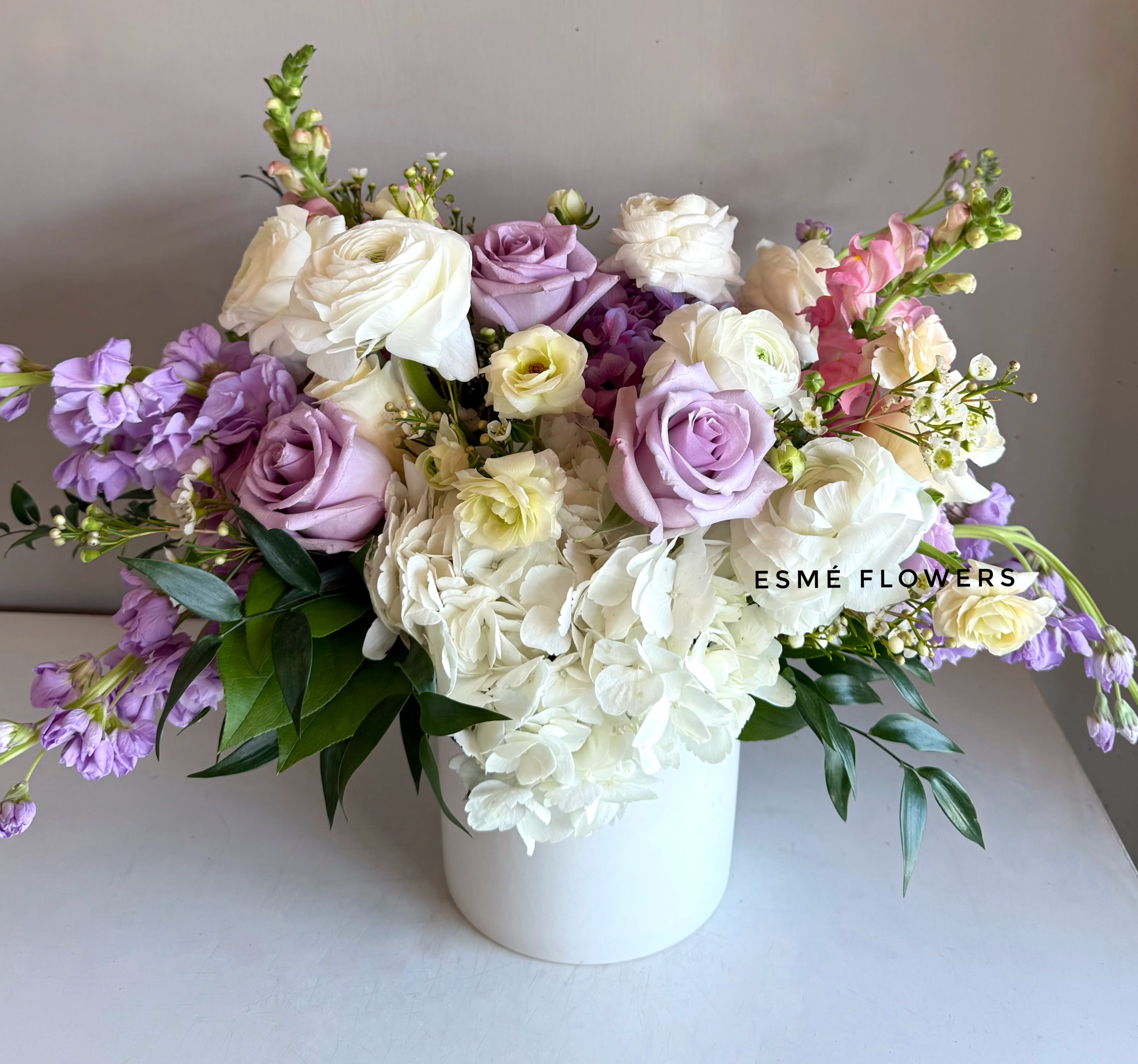 Fiora - A serene blend of soft lavender and creamy white blooms in a white ceramic vase. Fiora captures a sense of quiet elegance and gentle beauty.