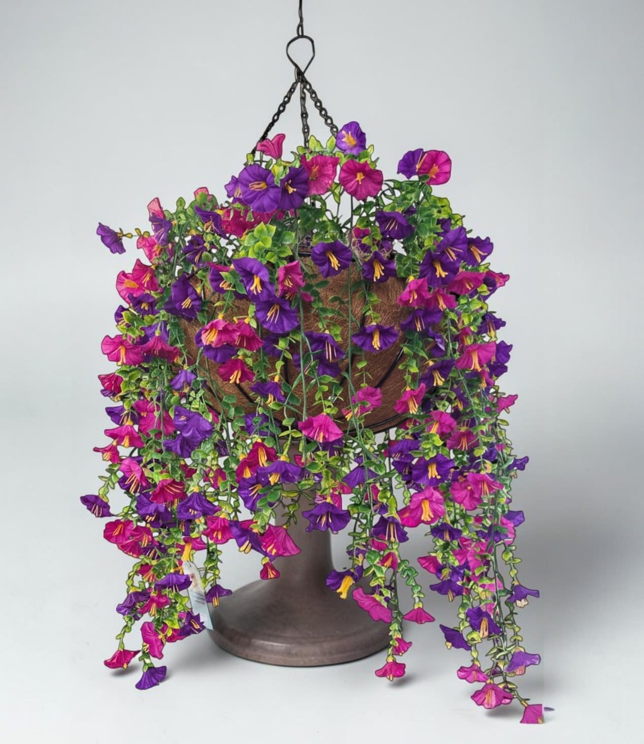 Violet Cascade by Art and Flowers - This hanging basket is a burst of saturated, joyful color — the kind of display that stops you mid‑walk because it feels so alive.    A lush cascade of blooms spills over the edges of the basket, dominated by rich purples, magentas, and hot pinks. The petals are velvety and full, with bright yellow centers that add a spark of warmth. The flowers trail gracefully, creating a soft waterfall effect, while the dense green foliage fills in the spaces with texture and depth. Suspended by simple black metal chains, the whole arrangement feels vibrant, abundant, and effortlessly eye‑catching — the kind of plant. T that thrives in sunlight and rewards you with nonstop color. This is a silk creation that is weather resistant. Other colors are available.