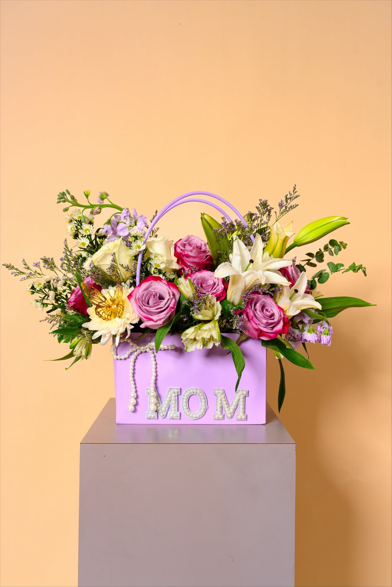 Lavender Grace for Mom - This elegant arrangement blends soft lavender tones with creamy whites and blush pinks, creating a calm and graceful expression of love. Delicate lilies and roses are beautifully framed in a pastel basket adorned with pearls and a golden butterfly. It’s a gentle, heartfelt tribute that honors a mother with timeless beauty and charm.