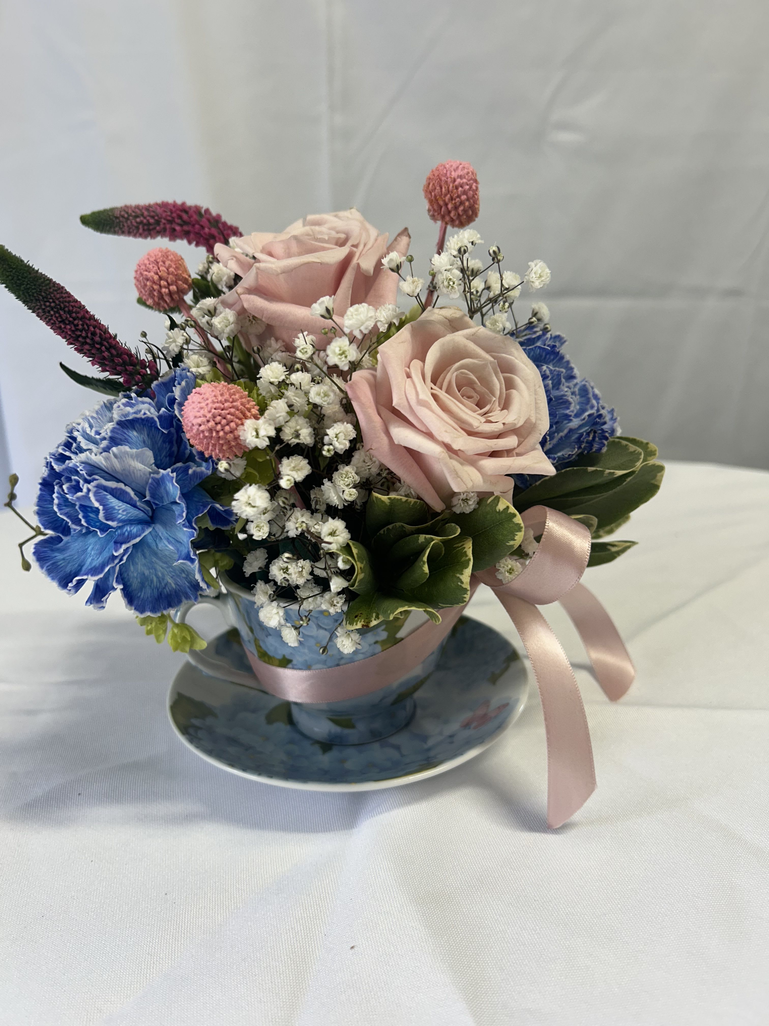 Pink roses and blue carnations arranged in a teacup with a ribbon
