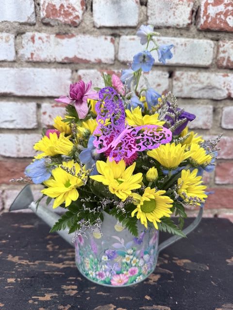 WATER ME WITH FLOWERS - A bright cottage-style arrangement brimming with yellow daisy-like blooms and pops of purple, blue, and pink, perfect as a birthday bouquet, thank-you gift, or just-because surprise. The Spring Floral Watering Can delivers an upbeat, playful impression that will make the person you're thinking of feel cheered and seen-ideal for celebrating everyday moments or bringing sunshine to a bedside or kitchen table.  This loose, rounded composition mixes saturated and pastel tones with small filler sprigs for texture, creating a lively, informal look. The flowers are gathered in a decorated tin bucket printed with floral artwork and tied with a decorative ribbon, giving the gift a charming, ready-to-display presentation. Sized to sit happily on a table or desk, it arrives arranged and gift-ready-no extra vase needed-so the recipient can enjoy the color and scent right away.