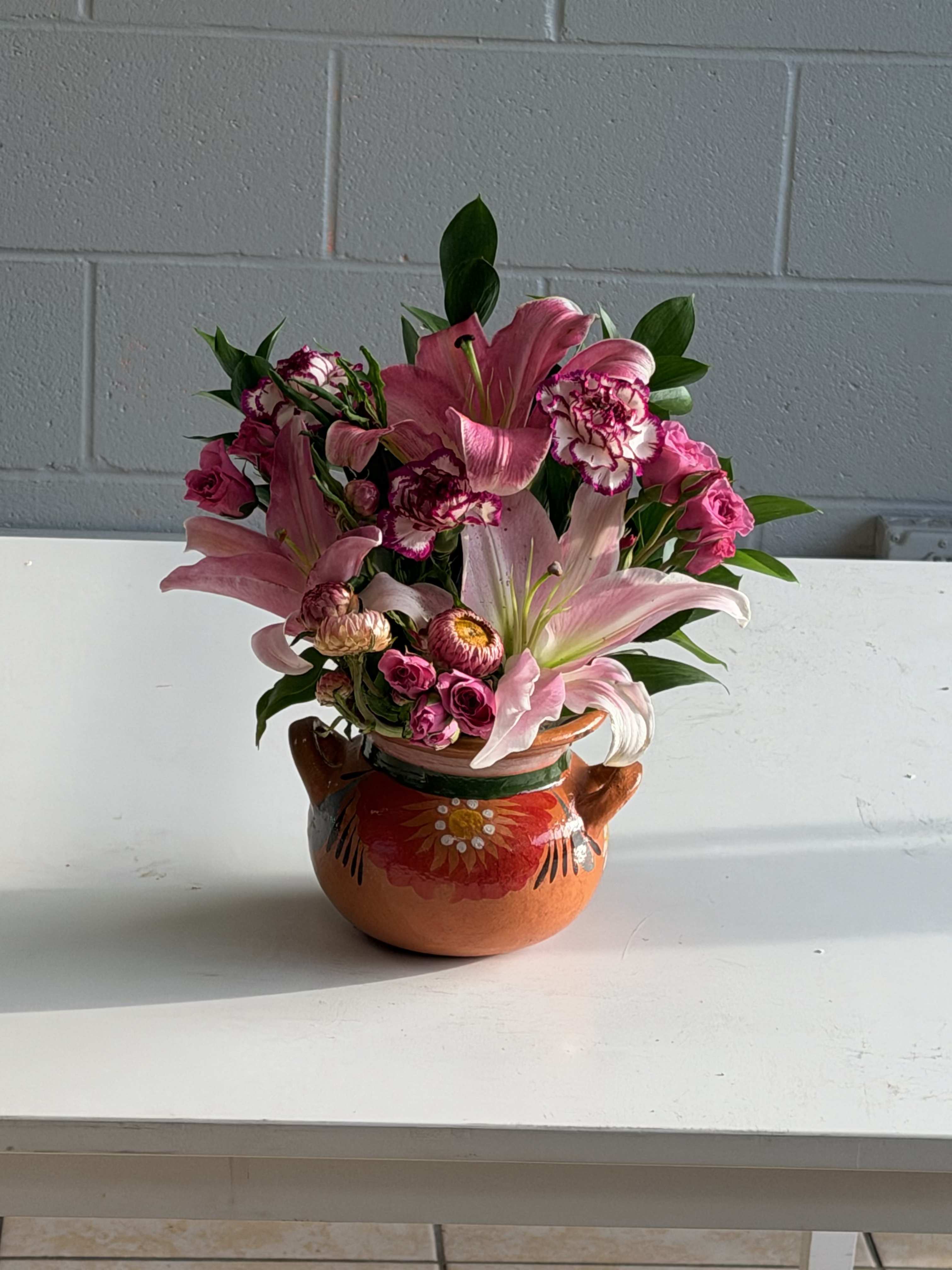 ollita de lirios - Bright pink lilies take center stage in this cheerful flower arrangement, a perfect birthday bouquet or thank-you gift that will make the person you're thinking of feel truly celebrated. The composition reads casual and handcrafted rather than formal, making it an ideal choice for spring occasions, a cheerful get-well surprise, or simply to say "thinking of you" with warmth.  Soft blush and brighter magenta carnations and small spray roses nestle around the lilies, with white accents and glossy green foliage for lively contrast. The blooms are arranged compactly in a low, rounded painted  vase (an orange clay pot with hand-painted floral details and side handles in some versions), giving the piece a rustic, handmade charm. The rounded silhouette and tight gather of flowers create a full, abundant look that feels intimate and feminine.  Give this to a friend, mom, or colleague who likes fresh, colorful arrangements-it's the kind of gift that reads joyful and personal. The sturdy ceramic or clay vessel arrives ready to display; keep water fresh and trim stems every few days to extend bloom life and keep the arrangement looking its best.