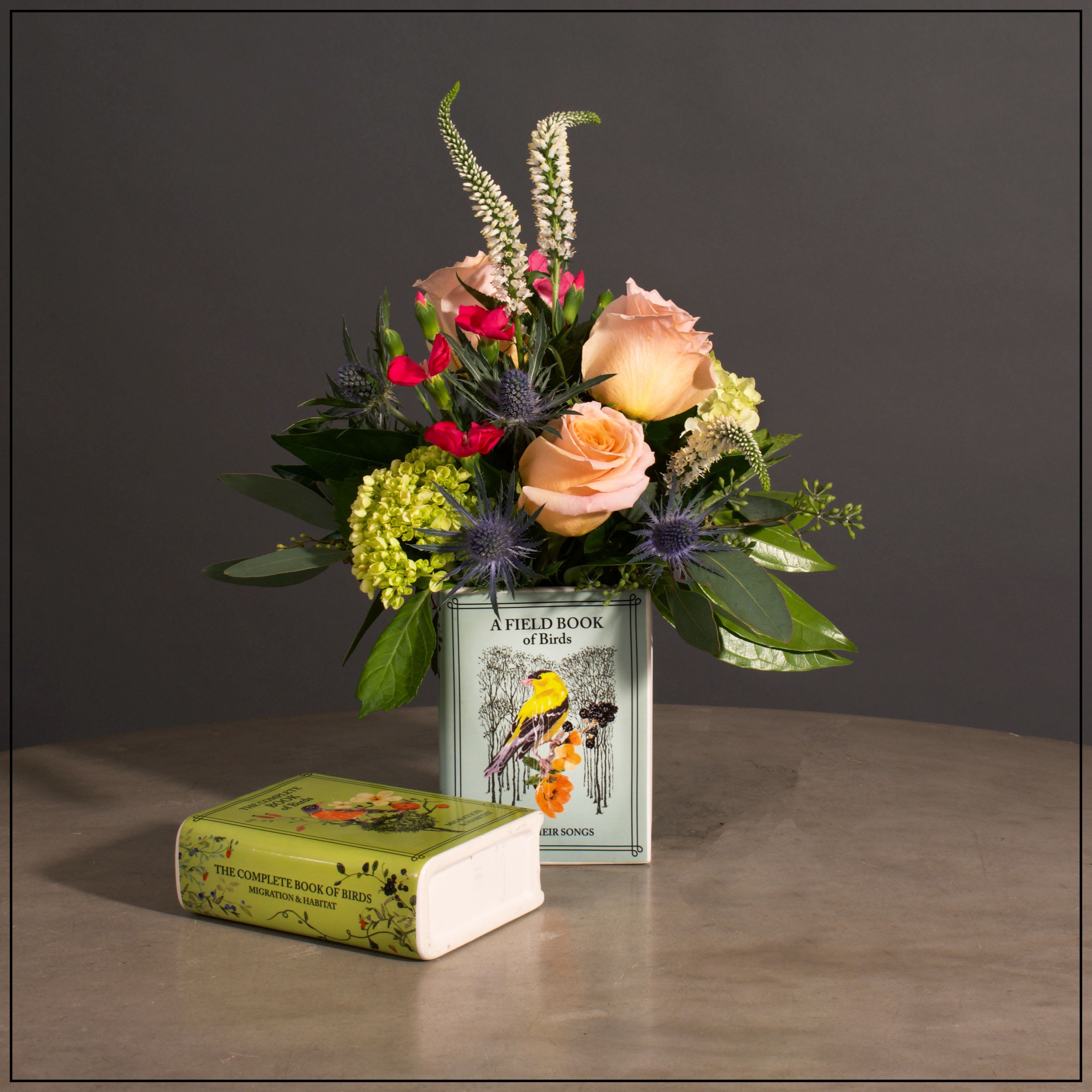 Bird Watch - Hydrangea, Roses, Veronica, Thistle and Solomio with Mixed Greens in a Unique Vase. The Vase is a Ceramic Recreation of a birdwatching Book with designs on Both Sides.  There Are 2 Assorted Styles (1 Blue and 1 Green; unless specified the designer will choose.)  Approximately 15" H x 10" W