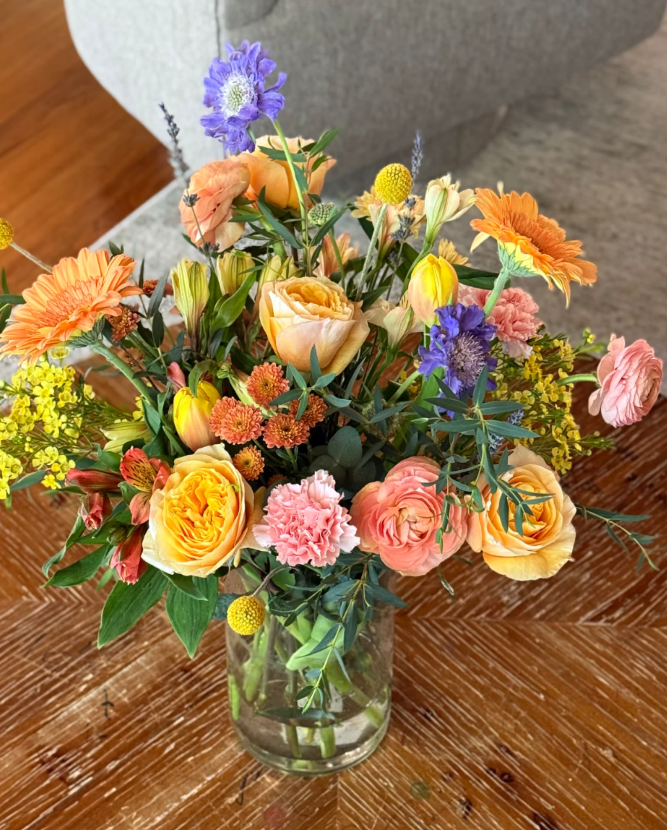 Willow Blossom - A lively, garden-style bouquet that greets the eye with warm orange and peach roses and sunny gerbera daisies-perfect as a birthday arrangement or a cheerful "just because" surprise. Willow Blossom's bright mix of yellow alstroemeria, pink carnations, soft ranunculus and tulip hints, plus purple accents, creates an uplifting, hand-gathered look that makes the recipient feel truly seen and celebrated.  Arranged loosely in a clear glass vase, the composition uses airy stems and small filler blooms to add texture, movement, and a fresh, layered silhouette that suits spring and summer moments. Large focal flowers anchor the design while smaller chrysanthemums and other seasonal touches keep it feeling abundant without fussy formality.  Send Willow Blossom to say thank you, brighten a get-well room, or mark Women's Day or Best Friends Day with joyful color. Vase-ready on delivery; refresh stems and change water every couple of days to keep blooms looking their best.