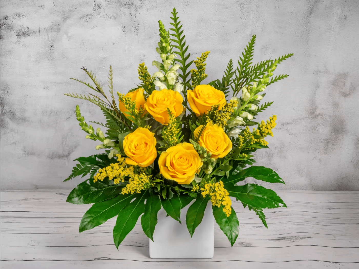 Yellow roses arranged in a white square vase with mixed greenery