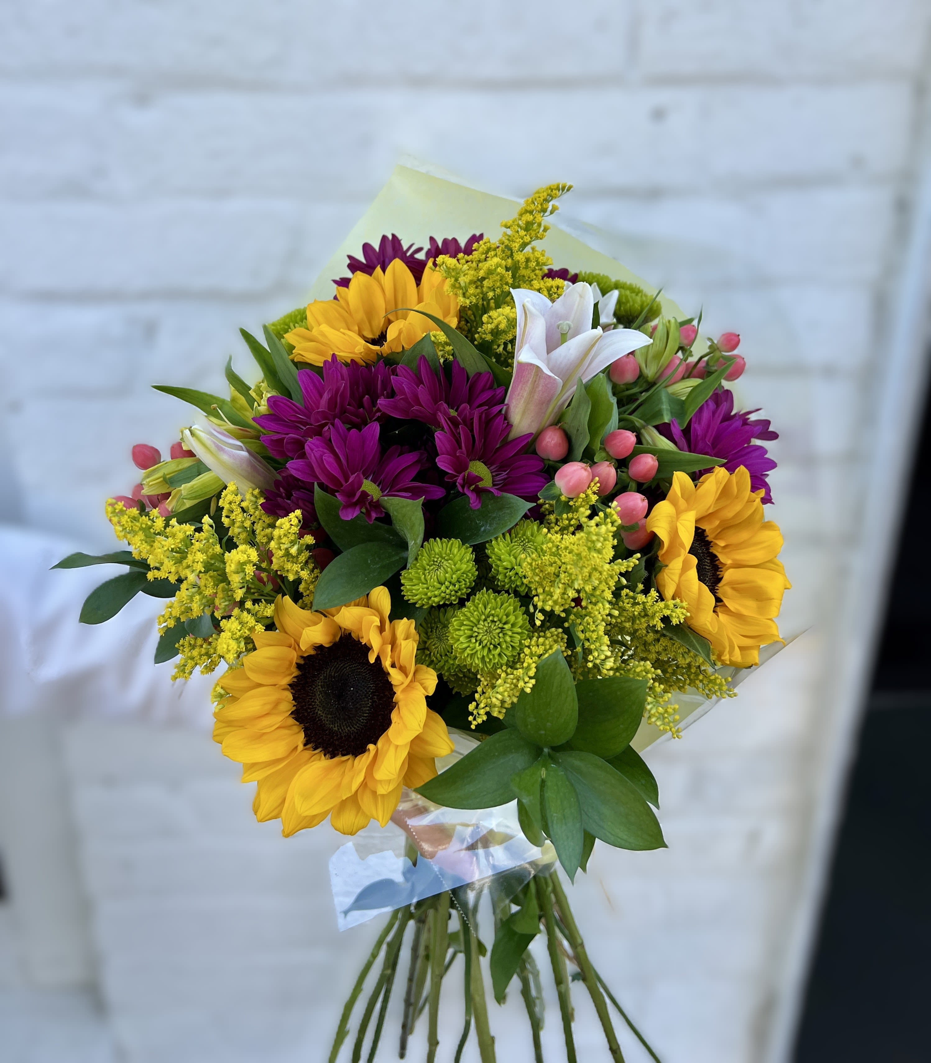 Sunflower Cheer - Bright, golden sunflowers steal the show in this sunflower bouquet built to brighten someone's day-ideal as a birthday bouquet, a cheerful get-well gift, or an everyday arrangement to say "I'm thinking of you." The warm yellow blooms paired with deep purple daisy-like flowers create an uplifting contrast that feels both lively and gently rustic, perfect for celebrating small moments or sending a confident, sunny message.  This hand-tied arrangement mixes sunflowers with white lilies, yellow filler blooms, green button chrysanthemums, and clusters of small pink berry accents for texture and dimension. Stems are gathered and finished in clear plastic wrap for a gift-ready presentation that keeps the bouquet tidy and easy to transport. Sized for tabletop display, it arrives ready to trim, place in water, and enjoy-she'll feel genuinely cheered the moment it's set out. NO VASE included.