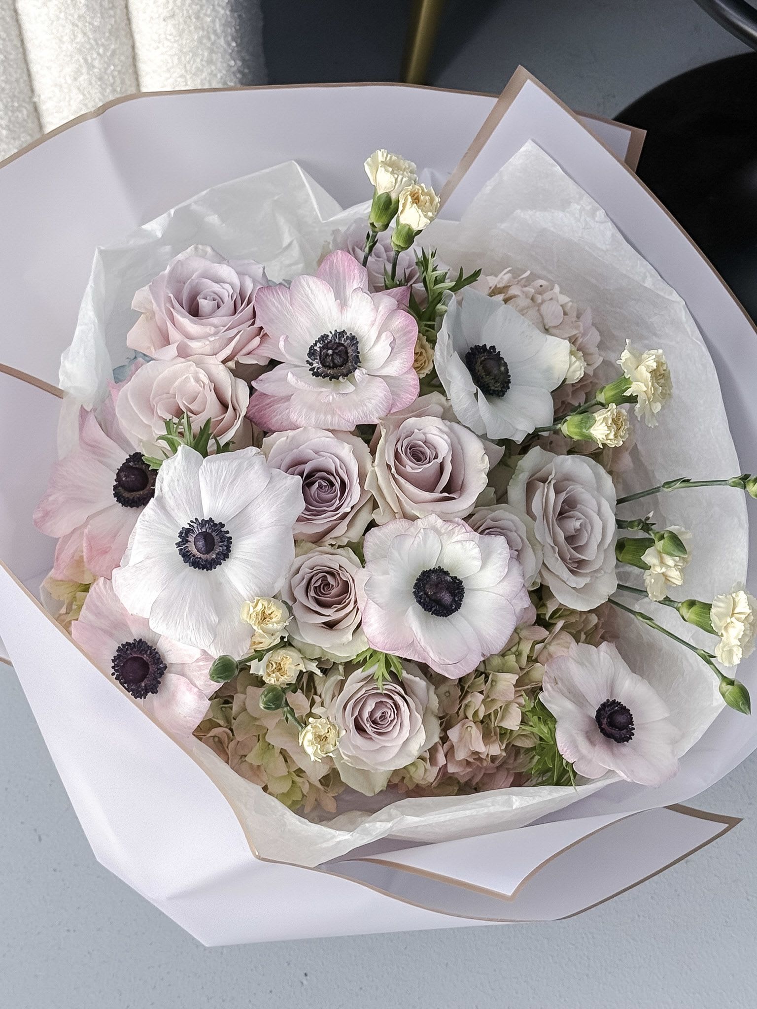 Midnight Blush - A soft, romantic composition with a touch of contrast, Midnight Blush blends delicate pastels with striking detail. Elegant black-and-white anemones take center stage, their bold hearts creating a captivating focal point among velvety quicksand roses in muted blush tones. Fluffy white hydrangeas form a cloud-like base, while mini spray cream carnations add texture and gentle movement throughout.  This bouquet feels modern yet timeless—perfect for expressing refined love, quiet elegance, or celebrating life’s most graceful moments.  Note: Anemones are naturally delicate and have a shorter vase life compared to other blooms. To enjoy them at their best, we recommend placing your bouquet in fresh water immediately and keeping it in a cool area away from direct sunlight.
