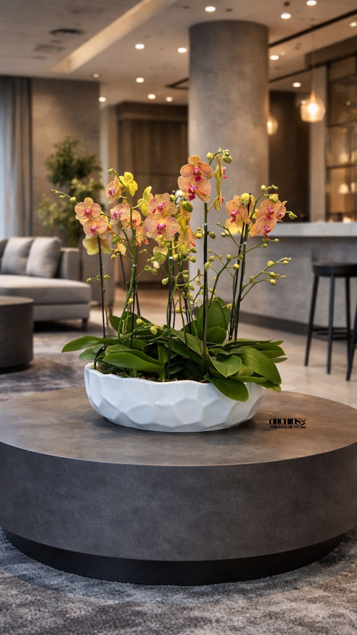 Low white bowl of yellow and pink orchids on a round gray table in a modern lobby.