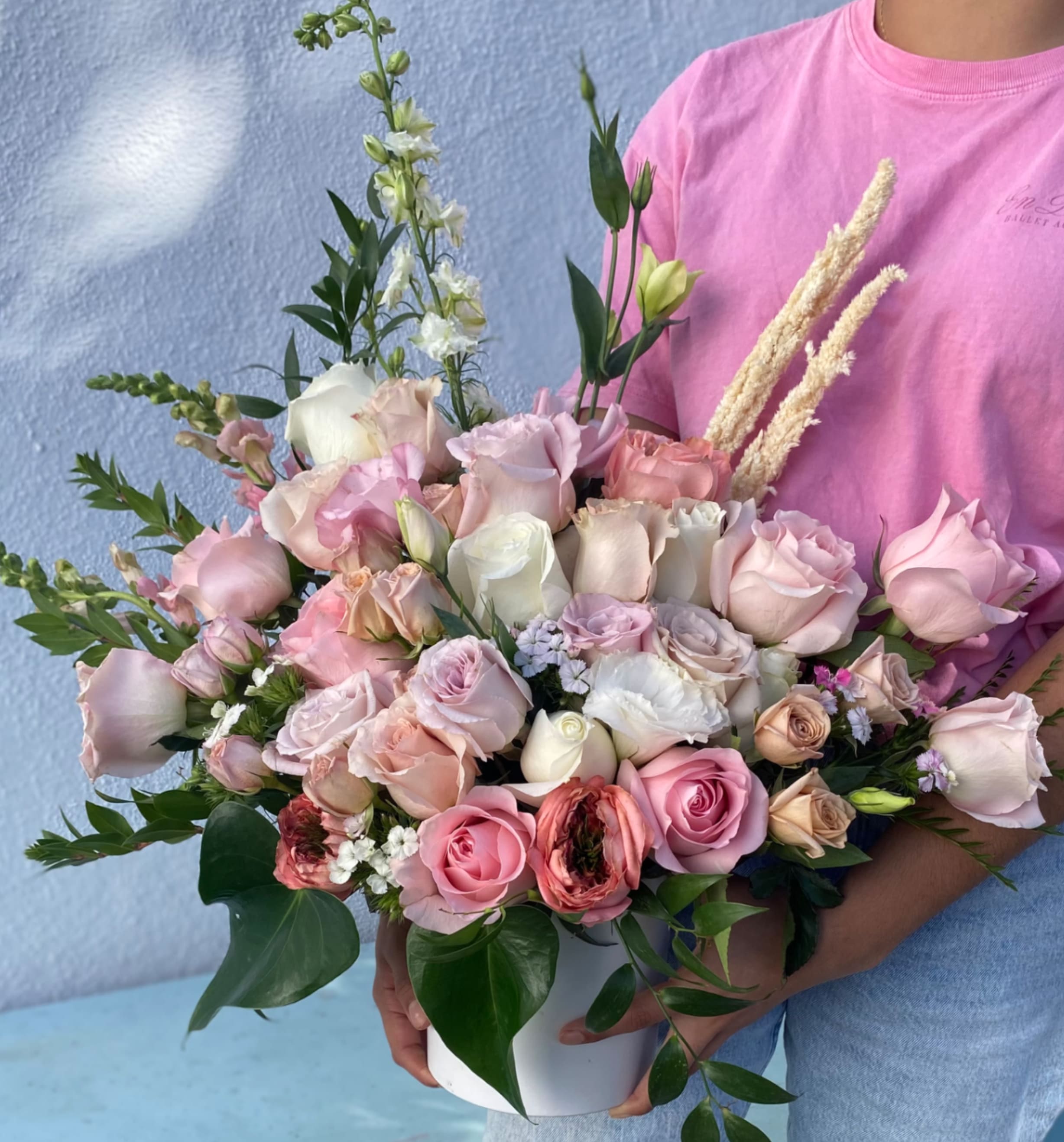 La Rosalia - Lush pastel roses and airy snapdragons combine in a garden-fresh bouquet that reads like a love letter in bloom - perfect as a birthday bouquet, anniversary flowers, or a Mother's Day gift that says you're thinking of her. This romantic arrangement of roses, lisianthus and snapdragons is styled in a hand-held, rounded shape and presented in a clean white container, so it arrives feeling both polished and genuinely hand-tied for occasions of love and gratitude.  Soft pink, blush, cream, peach and touches of green and purple mingle across the design: abundant roses form the romantic core, lisianthus lend delicate buds and ruffled texture, and tall snapdragon spikes add height and movement. Broad glossy foliage frames the base for a full, garden-like silhouette that feels both substantial and tender.  Whether you're celebrating a wedding, saying thank you, marking an anniversary, or surprising someone just because, she'll feel noticed and cherished when this bouquet arrives. Arrange on a table or mantel as-is in its white container for immediate impact; to keep blooms looking their best, refresh the water and trim stems every few days.