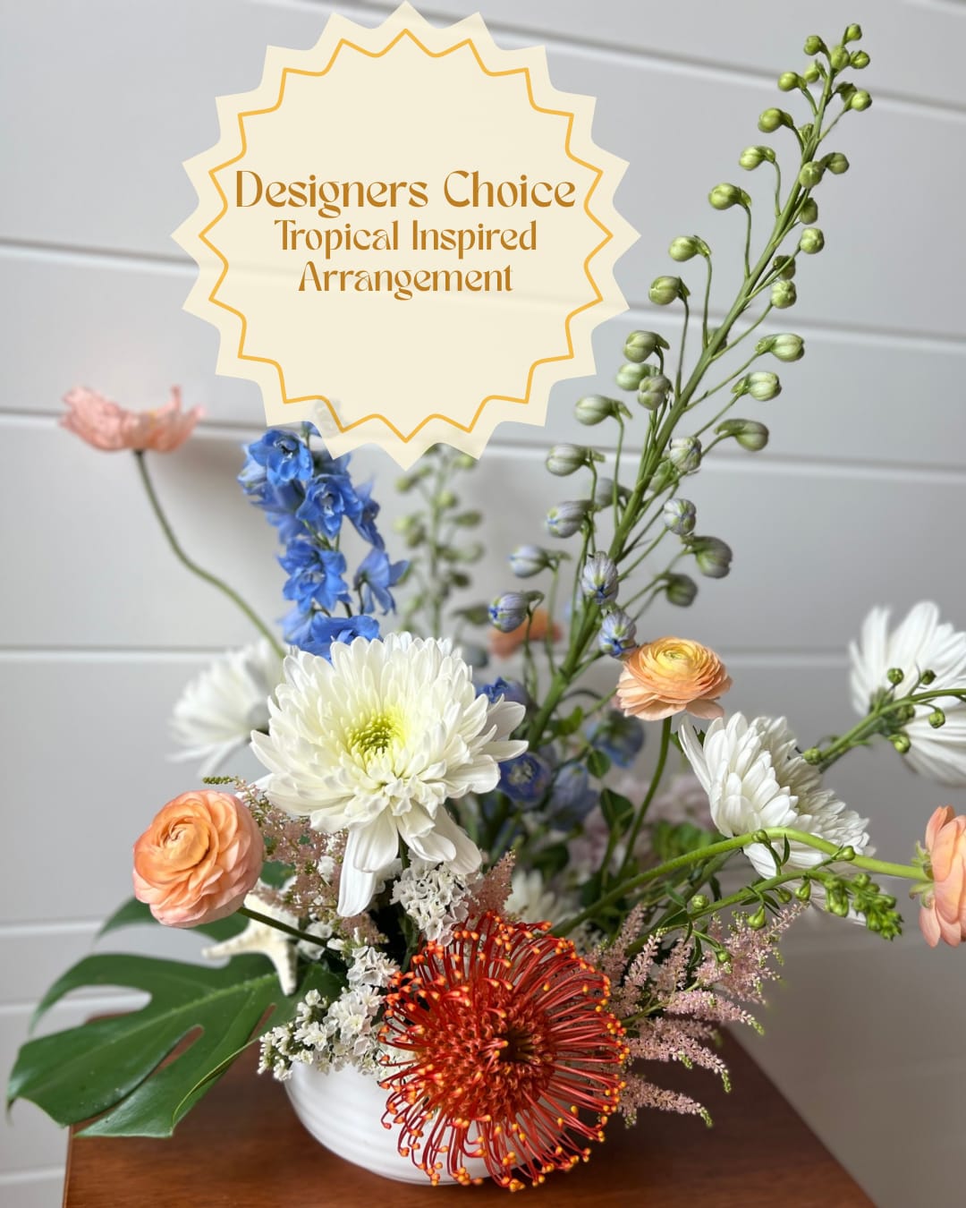 Designers Choice- Tropical Inspired Arrangement - A fresh, tropical-inspired flower arrangement that feels both bright and relaxed-designed as a cheerful birthday bouquet, congratulations gift, or everyday thank-you arrangement. The airy, layered silhouette and playful color pops make the recipient feel celebrated and noticed.  This designers-choice arrangement is built in a simple white ceramic vase and features tall branching stems for height, lower clustered blooms for fullness, and a broad tropical leaf at the base for contrast. Expect a mix of seasonal blooms complementing flowers in complementary tones for adding a fresh, slightly exotic touch to a home or office.  A lovely option when you want someone to feel uplifted and seen, whether for a birthday, promotion, or just because. Standard sizing will create the layered, airy profile shown; place in a cool spot away from direct heat and refresh the water every few days to keep blooms looking their best.