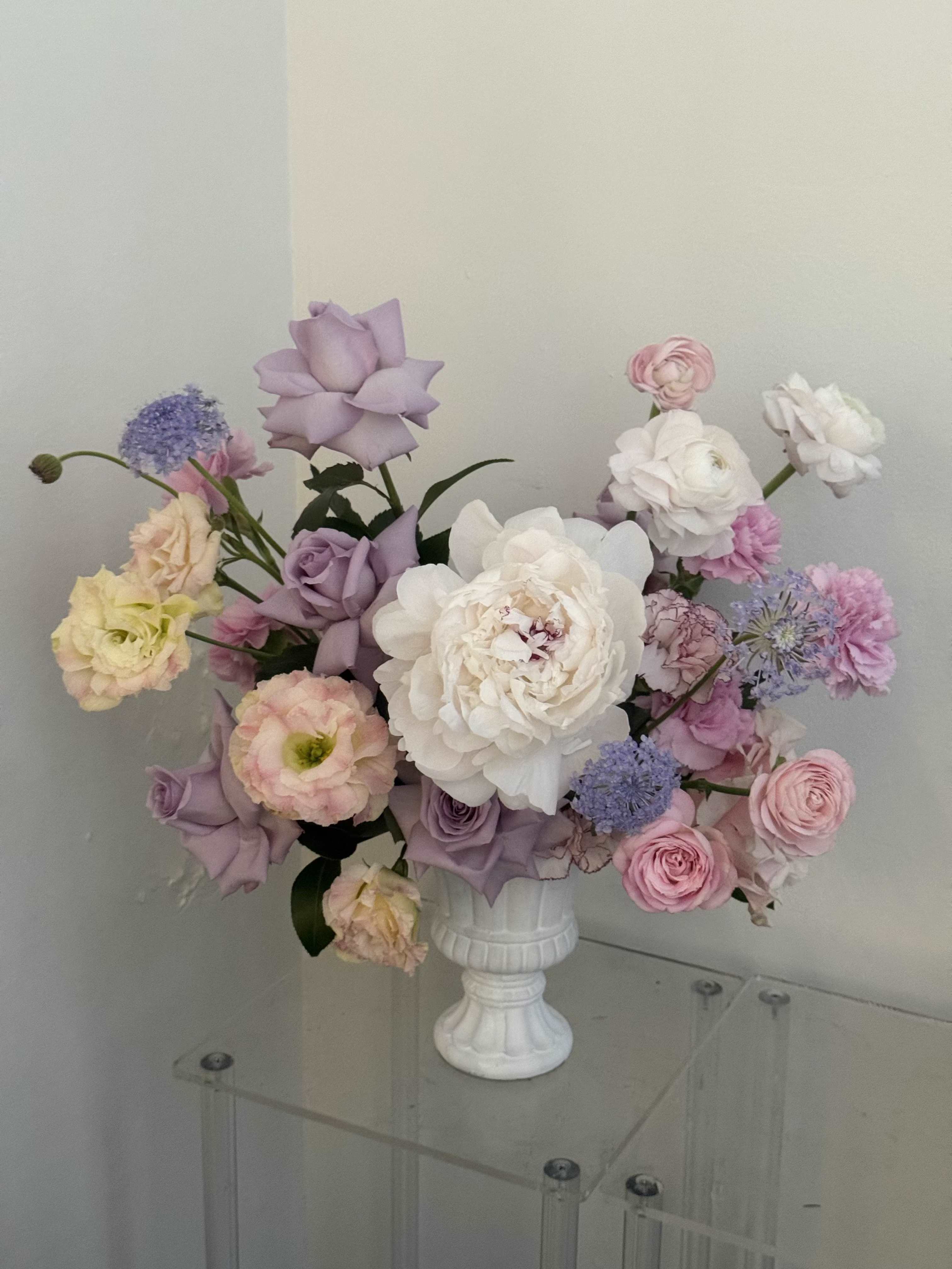 Blush Garden - Lavender and blush tones mingle like a spring garden in bloom-this pedestal arrangement of ranunculus, peonies, roses, lisianthus, and carnations is a soft, romantic gift for Mother's Day, an anniversary, or to welcome a new baby. The rounded, garden-style composition and airy pastel palette-pink, lavender, and touches of pale peach-creates an immediate sense of tenderness so the person you're thinking of will feel cherished and seen.