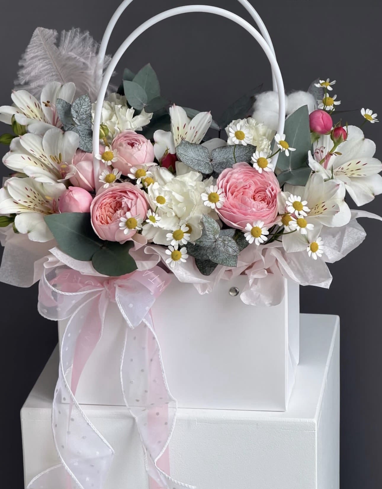 Tote of Garden Blooms - Soft pink roses and pale carnations mingle with white alstroemeria and dainty daisy-like blooms in this Tote of Garden Blooms-an airy, rounded flower arrangement perfect for birthdays, anniversaries, or Mother's Day. The romantic, feminine palette of pinks, whites, and muted greens is accented by silvery foliage for gentle contrast, creating the kind of bouquet that makes the person you're thinking of feel truly cherished.  Arranged in a white handled gift box and finished with a sheer pink ribbon and white tulle, this design reads as both elegant and effortlessly casual-ideal for sending warm wishes, saying "thank you," or marking a special milestone. The compact, tote-style presentation is easy to carry and display on a table or bedside, and its soft textures and pastel hues pair beautifully with delicate stationery or a handwritten note.