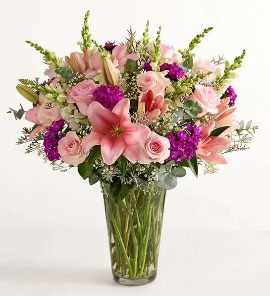 Marvelous Mom  - Soft pink lilies and pale blush roses take center stage in this romantic bouquet, making it an ideal birthday, anniversary, or Mother's Day arrangement. Vivid purple clusters and airy white accents weave through the composition to create a lush, celebratory feel-the kind of gift that makes the person you're thinking of feel truly seen and cherished.  The bouquet is arranged in a tall clear glass vase so long stems and fresh water are visible, giving a garden-fresh, hand-tied look. Large focal lilies and roses are balanced by dense purple filler blooms, delicate white accents, and soft, eucalyptus-like green foliage for texture and movement. The palette of pink, purple, white, and green reads feminine and abundant without being fussy.  Perfect for marking milestones or simply saying "I'm thinking of you," this arrangement arrives ready to display and enjoy. For best longevity, keep water fresh and trim stems every few days so the lilies and roses stay vibrant throughout the celebration.