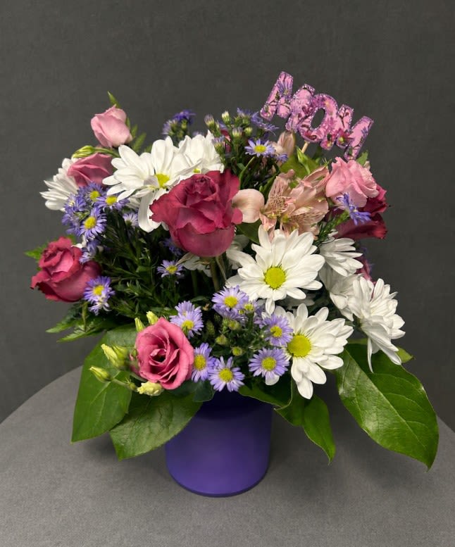 Mom’s Treasures - Pink roses, white daisies and purple blooms arranged in a compact bundle atop a purple vase, framed with glossy leaves and topped with a pink "Mom" topper-cheerful Mother's Day gift. 