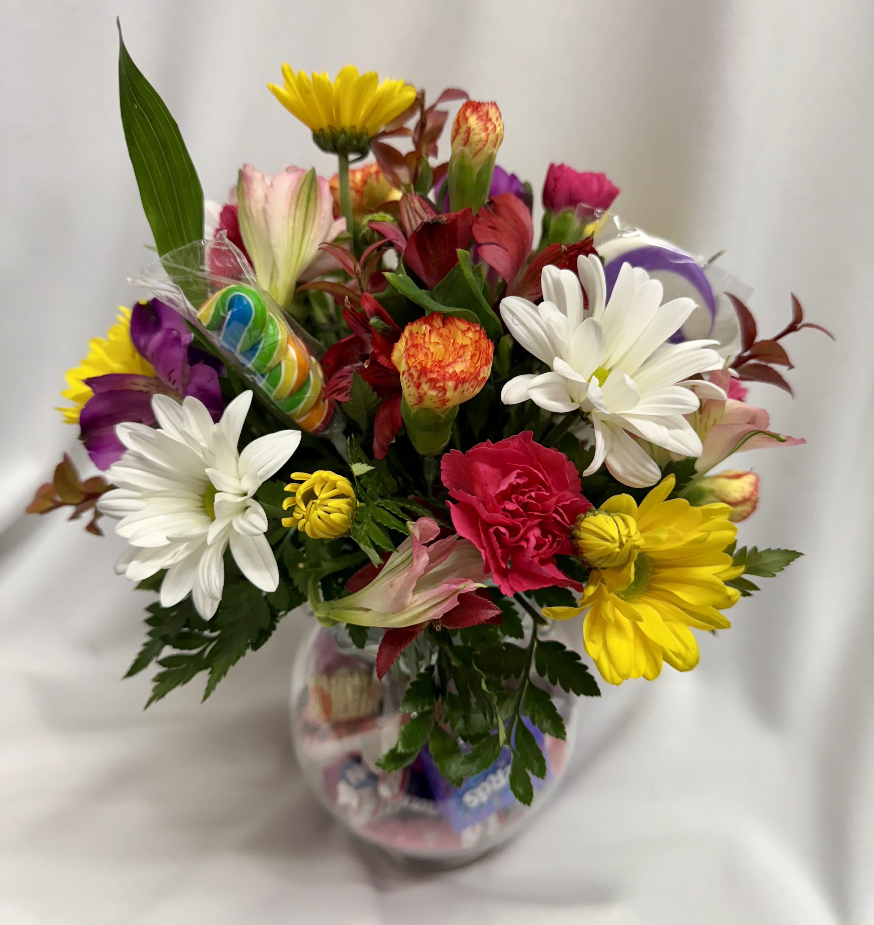 Sweet Artistry - A cheerful mixed-color flower arrangement, making it a joyful choice for a birthday bouquet, Mother's Day gift, or an Easter centerpiece. Sweet Artistry brings an upbeat, lighthearted mood that says "you're celebrated" to the person you're thinking of-perfect for graduations, get-well wishes, just-because surprises, or a romantic spring gesture.  The bowl-style clear glass vase holds a clustered mix of alstroemeria, roses, daisies, carnations, chrysanthemums and other small filler blooms in lavender, yellow, pink and green tones. Textural contrasts-from soft rose petals to crisp daisy faces-give the arrangement visual depth, while assorted wrapped candies packed at the base add a playful, two-gift surprise and suckers poked in with the flowers. The compact design works well on tabletops and desks and arrives ready to display.  This arrangement is ideal for brightening a mom's day, treating a graduate, cheering a patient, or saying thank-you to a favorite coworker during Admin Professionals' Week or Boss's Day. If you have a candy preference, include it in the special instructions at checkout so we can personalize the sweet touch.
