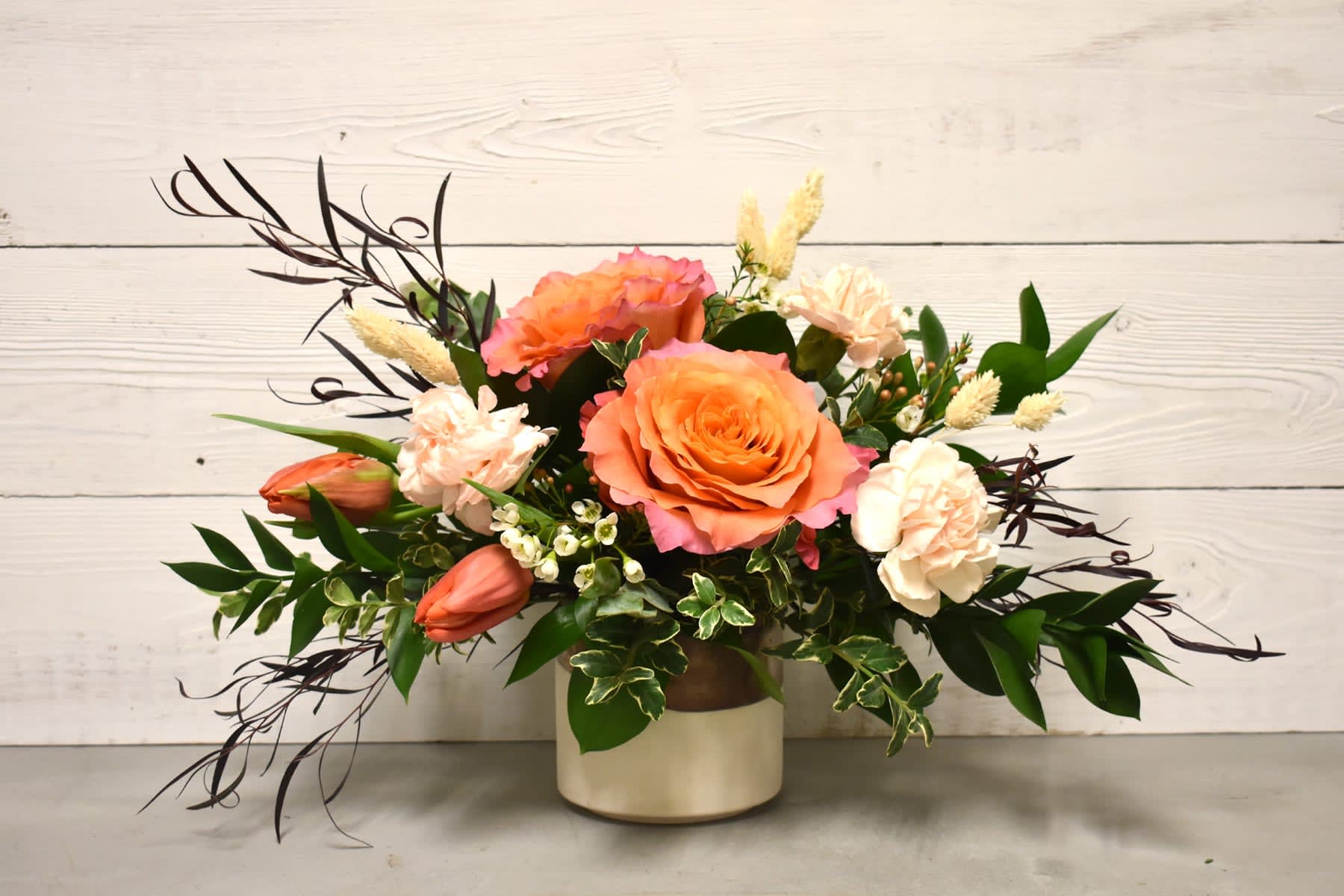 Low arrangement of peach roses, blush carnations, and orange tulips in a cream ceramic vase