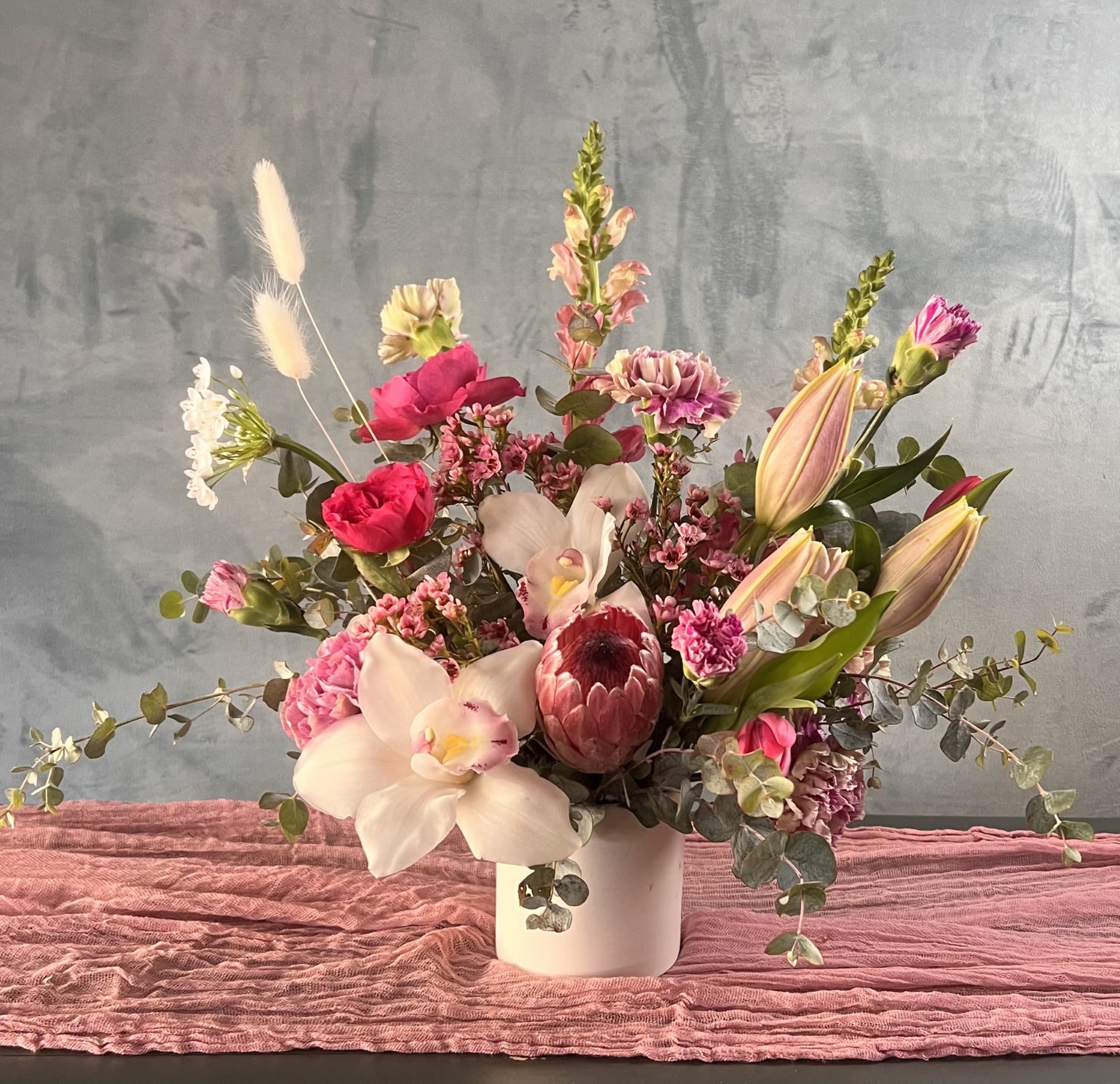 Nora - This structured floral arrangement is designed in refined shades of pink and white, featuring striking protea and elegant orchids. A composition that blends clean lines with sculptural elements, creating a modern, elevated design with strong visual impact.  Each arrangement is thoughtfully handcrafted using premium seasonal blooms, selected for their form, texture, and longevity. The combination of protea and orchids brings a unique balance of boldness and softness, resulting in a design that feels both contemporary and sophisticated.