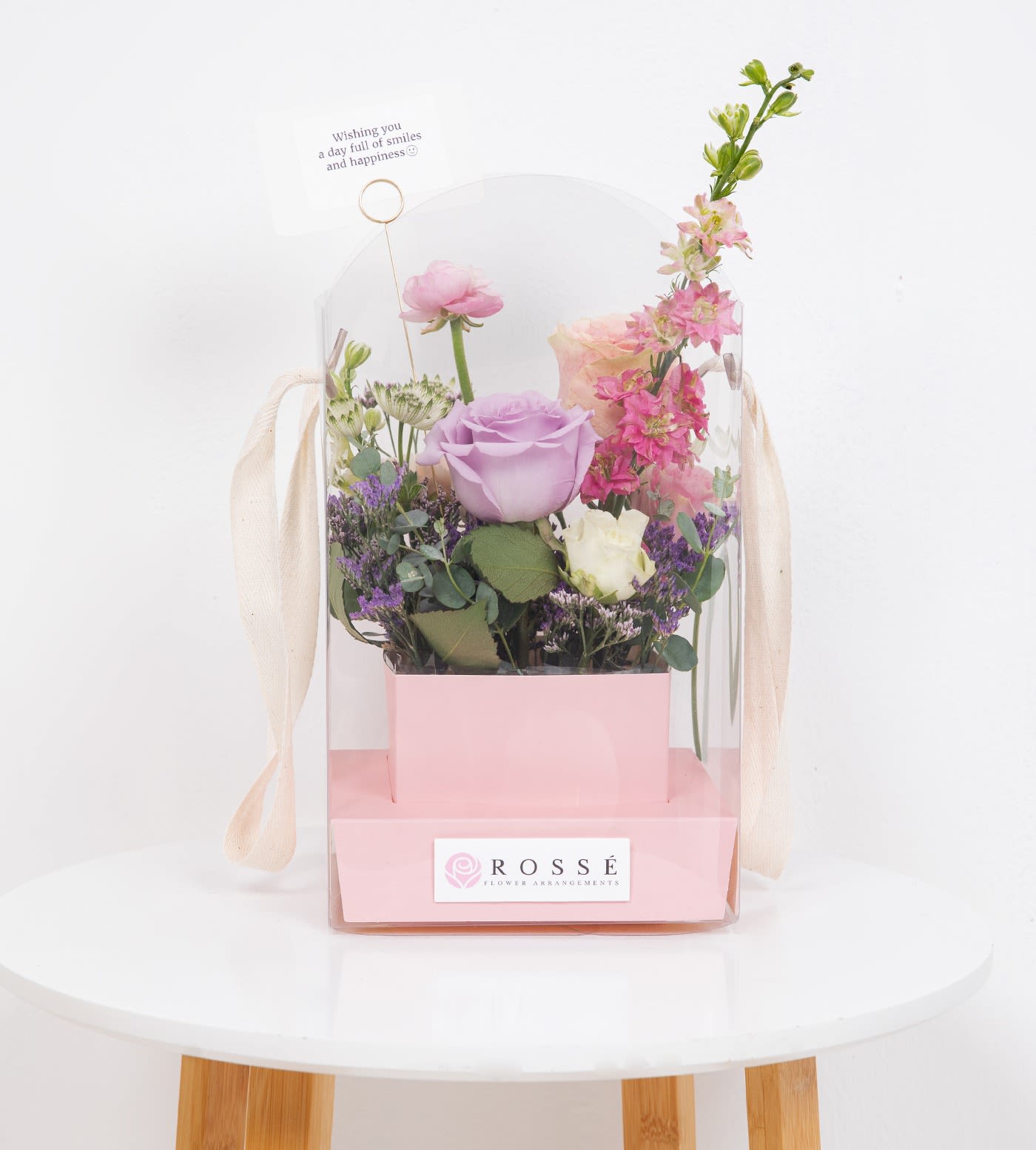 Pastel pink and purple floral arrangement in a pink box with clear cover and small greeting card.